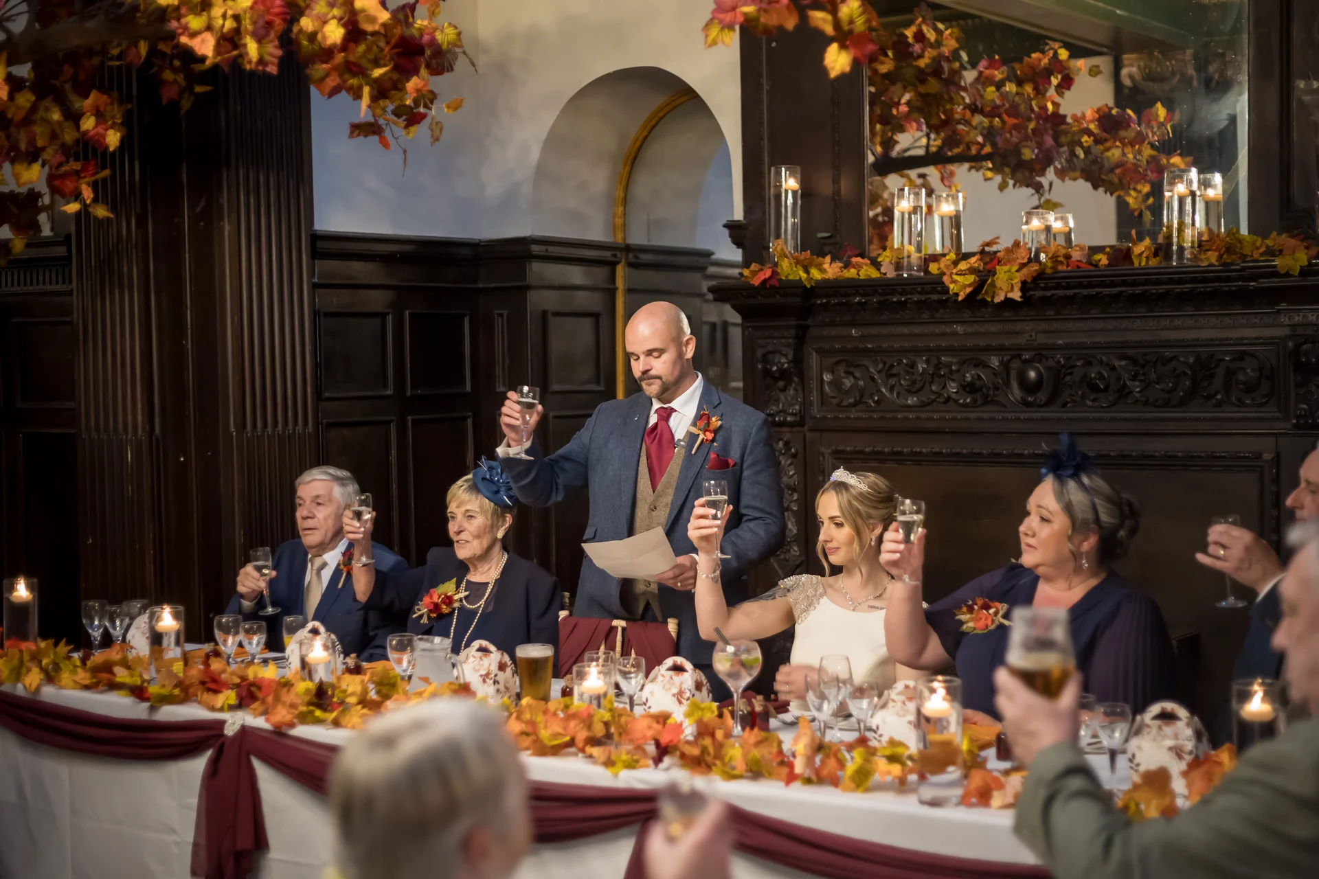 A groom stands at the top table during wedding speeches, holding a glass of champagne to toast while family members and guests raise their glasses in celebration amidst autumnal decor.