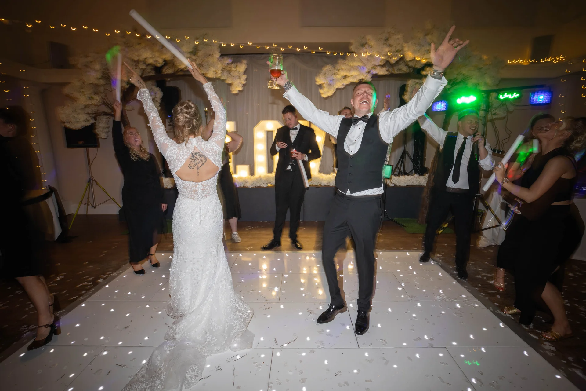 A joyful bride and groom celebrate on a glowing dance floor with cheering guests raising glow sticks during their evening wedding reception.