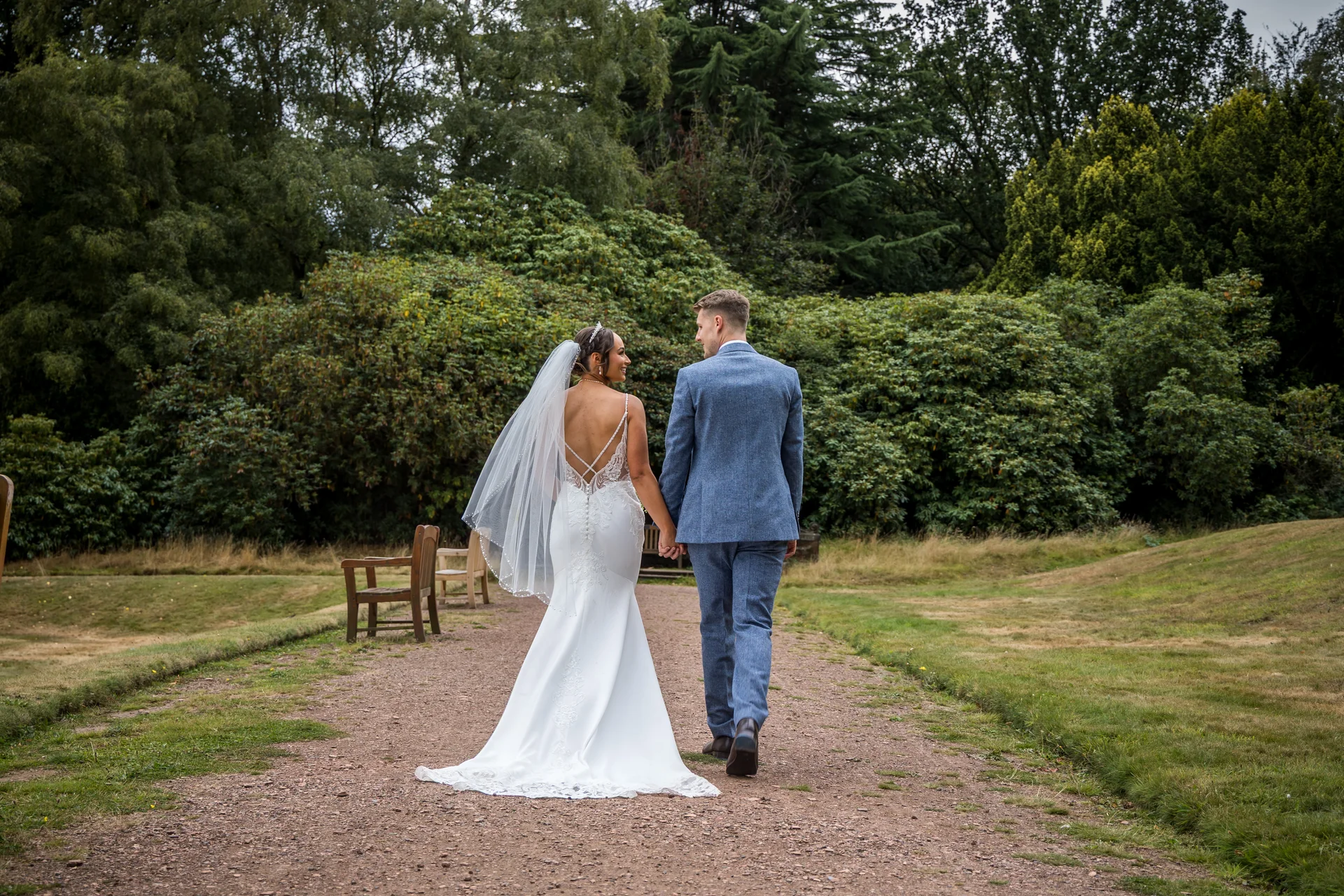 Newlyweds walking hand in hand along a gravel path in a lush green woodland, with the bride looking back over her shoulder.