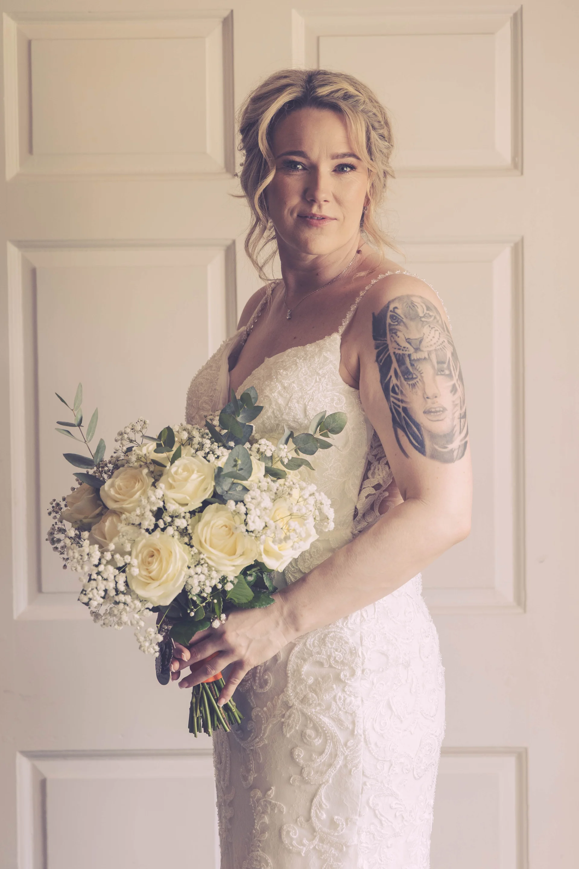 A stunning bridal portrait of a blonde bride with a textured bun, wearing a beaded lace gown with a V-neckline, showcasing a detailed arm tattoo while holding a hand-tied bouquet of white roses and gypsophila against a panelled white door.
