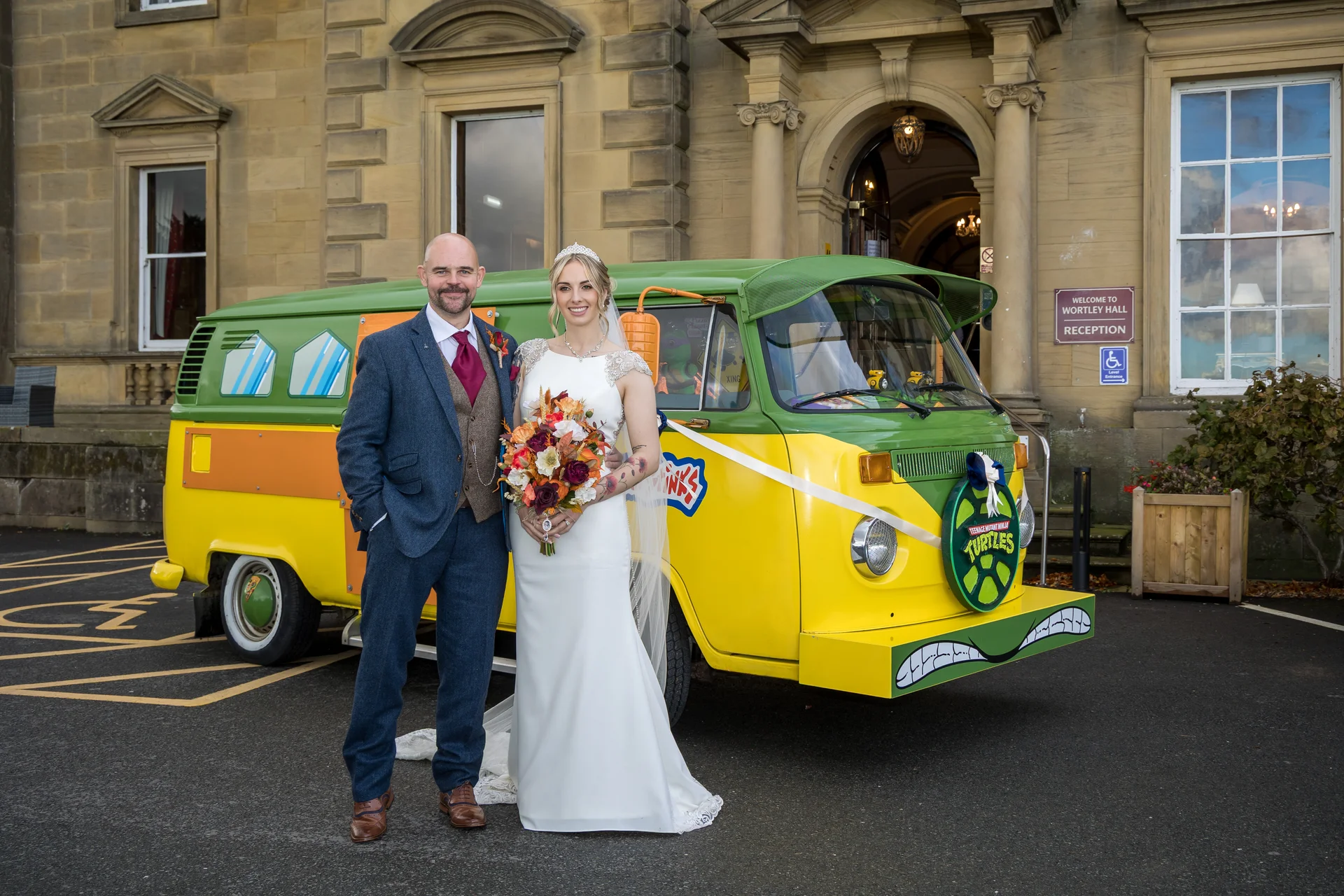 A bride and groom pose happily together in front of a grand stone manor house next to a brightly decorated green and yellow campervan.