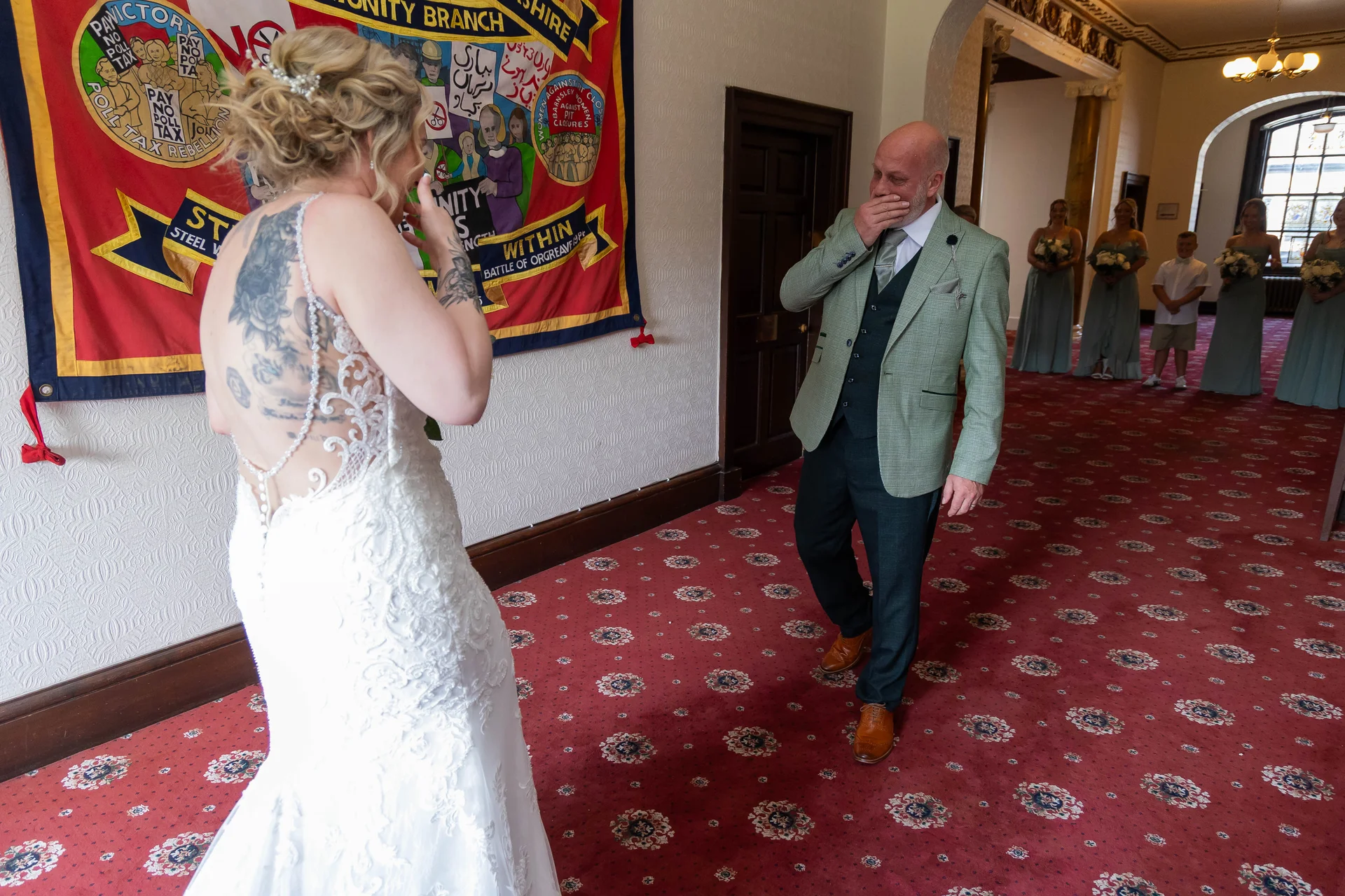An emotional groom is brought to tears as he catches a first look at his tattooed bride in her lace wedding gown inside a hotel hallway.