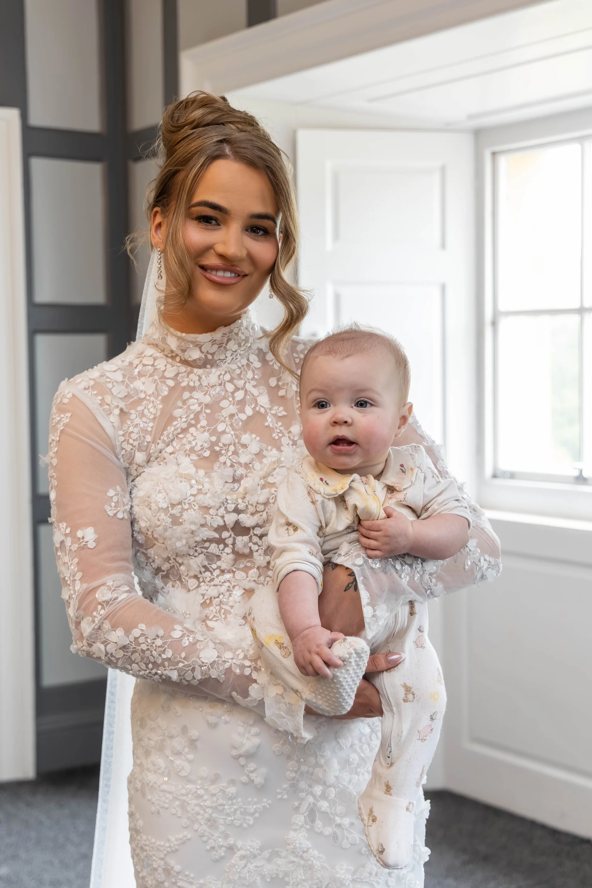A smiling bride wearing a high-necked, lace-apportioned long-sleeve gown gently holds a baby in a printed babygrow indoors near a bright window.