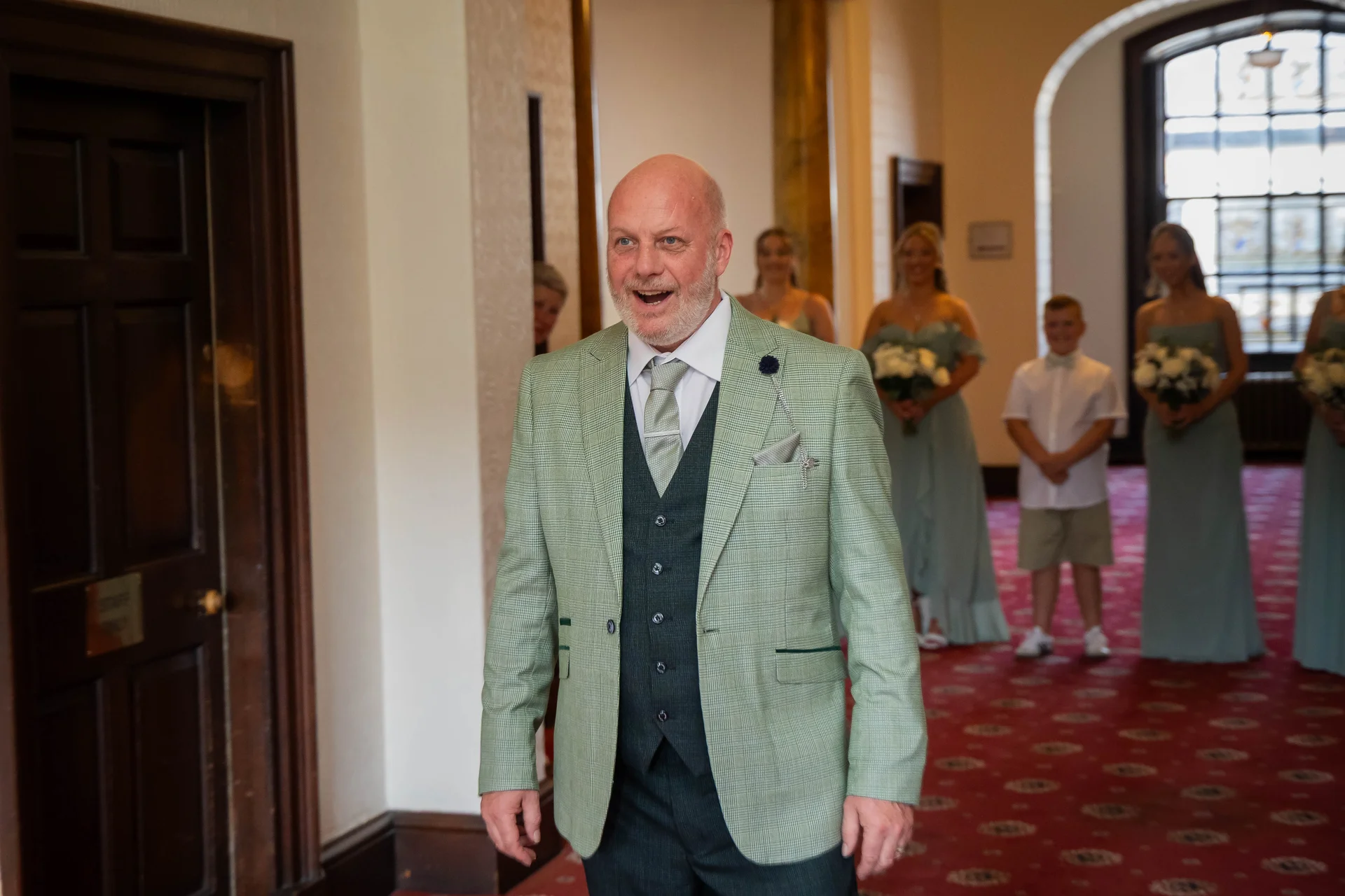 An excited groom in a patterned green jacket and dark waistcoat reacts with a joyful expression while waiting inside a hallway with family and wedding party members behind him.