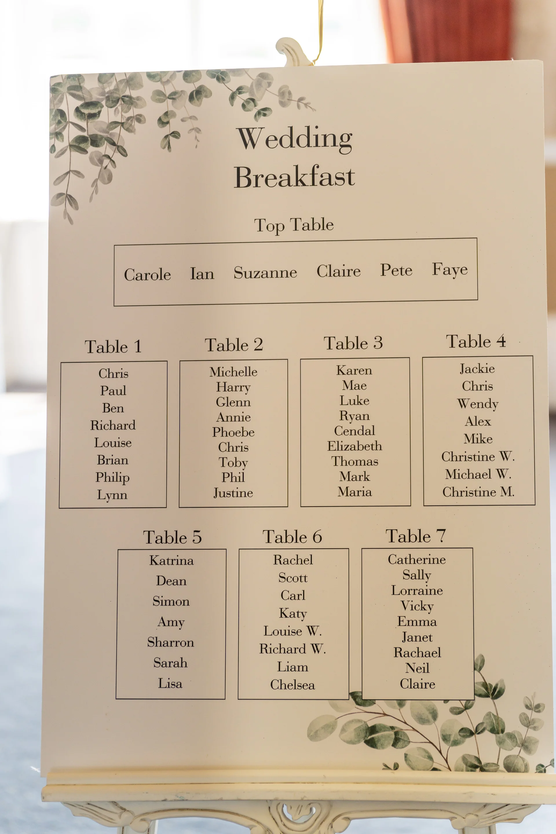 An elegant wedding breakfast seating plan displayed on an easel features clean typography adorned with delicate eucalyptus foliage illustrations.