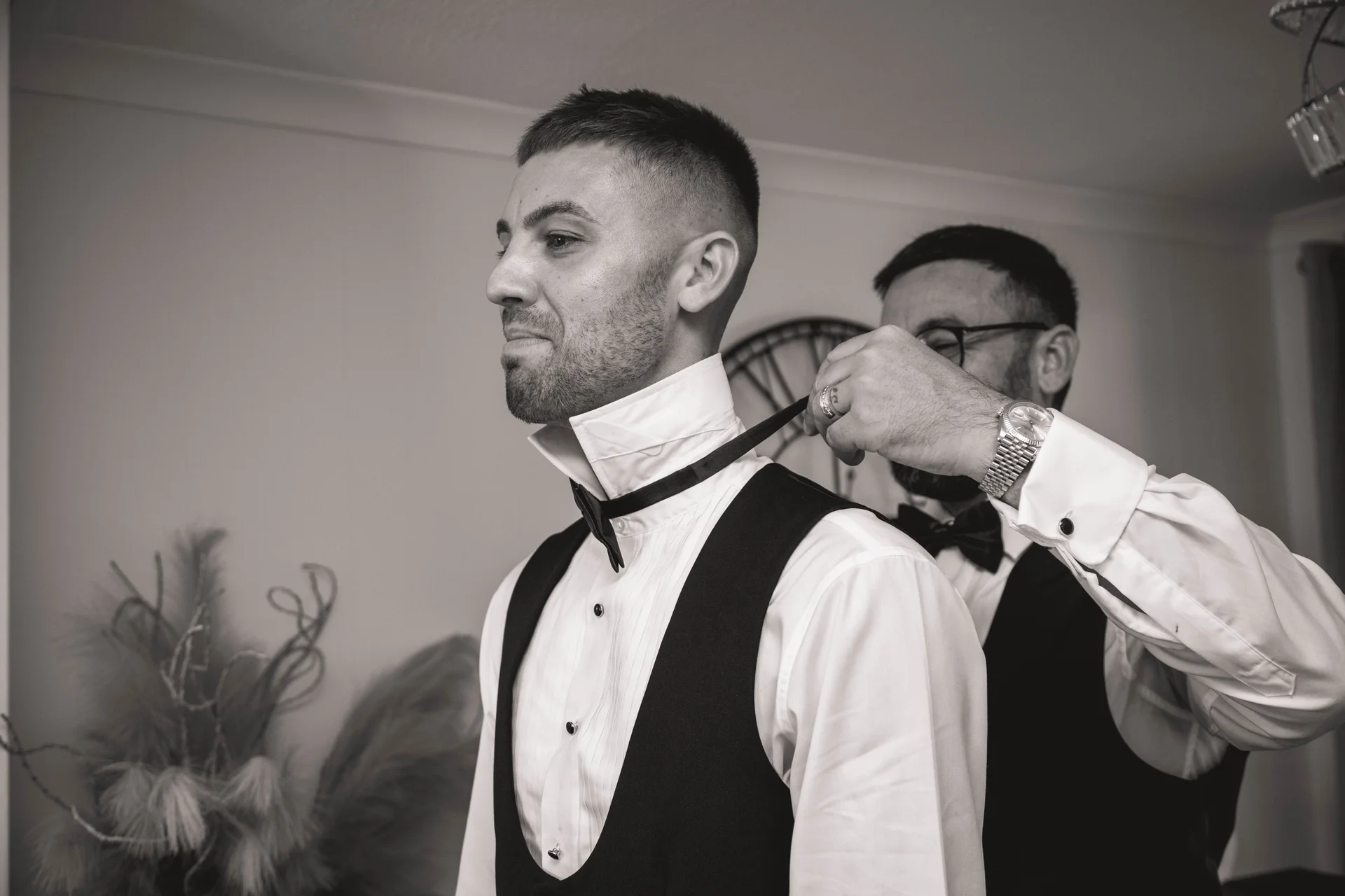 A groom in a white shirt and waistcoat gets his bow tie adjusted by a best man during morning preparations.