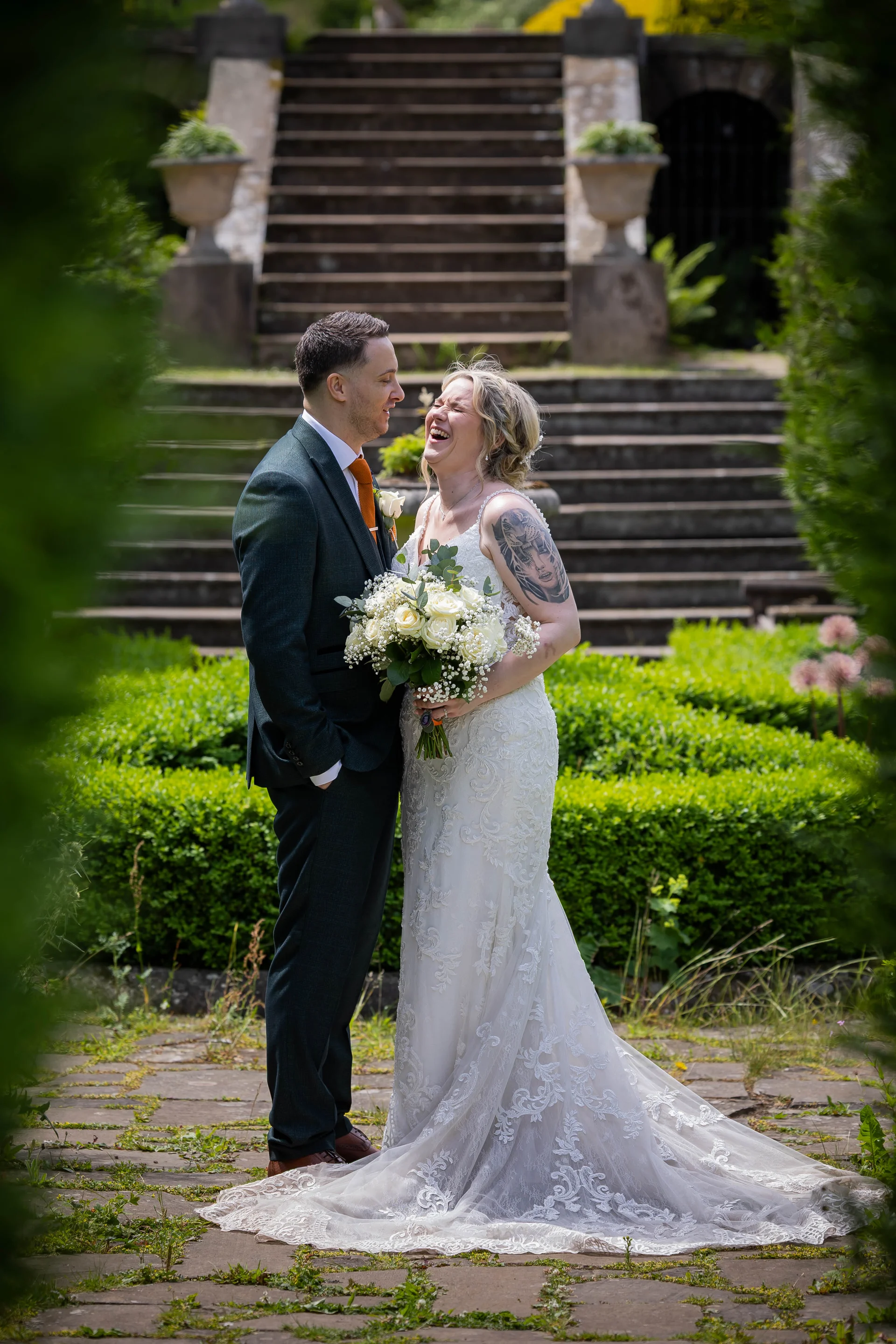 A newlywed couple laughing joyfully together in a manicured garden with stone steps in the background, the bride holding a white rose bouquet and showcasing arm tattoos while wearing a lace gown.