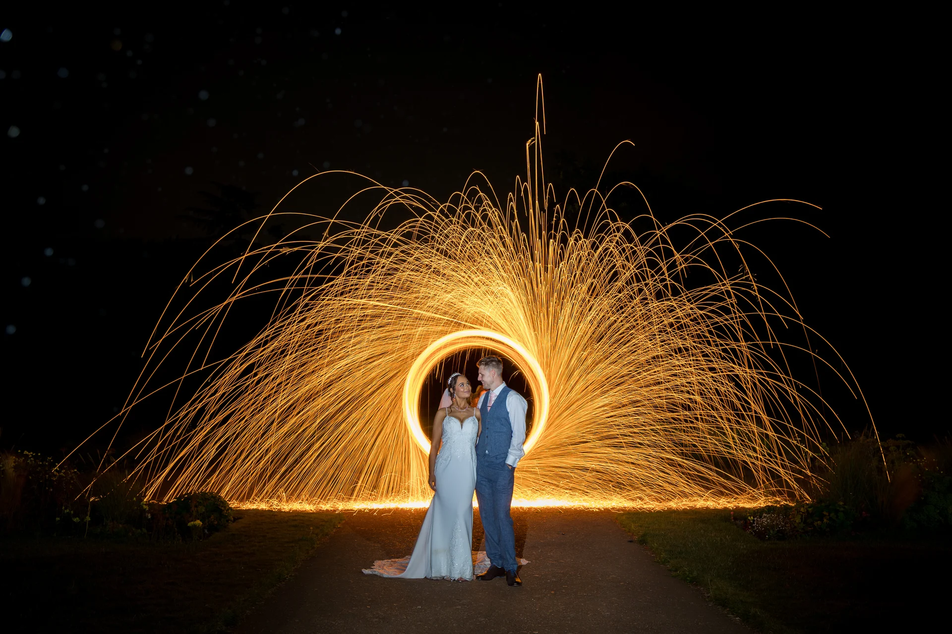 A newlywed couple stands together on a path at night, framed by a vibrant, circular arc of fire from a glowing sparkler light painting.