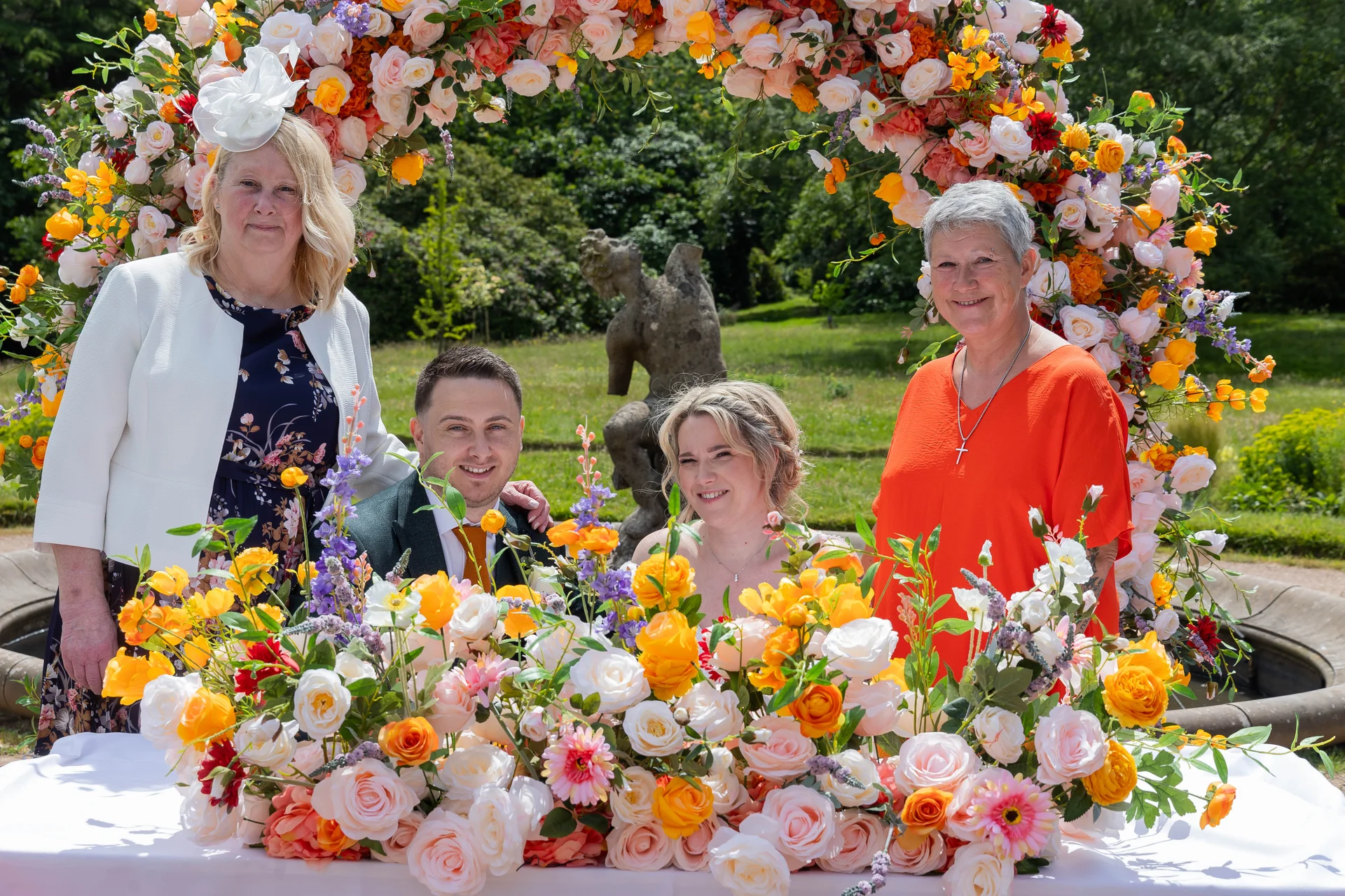 A smiling bride and groom pose outdoors with family members in front of a large, vibrant floral arch adorned with pink and orange roses in a manicured garden.