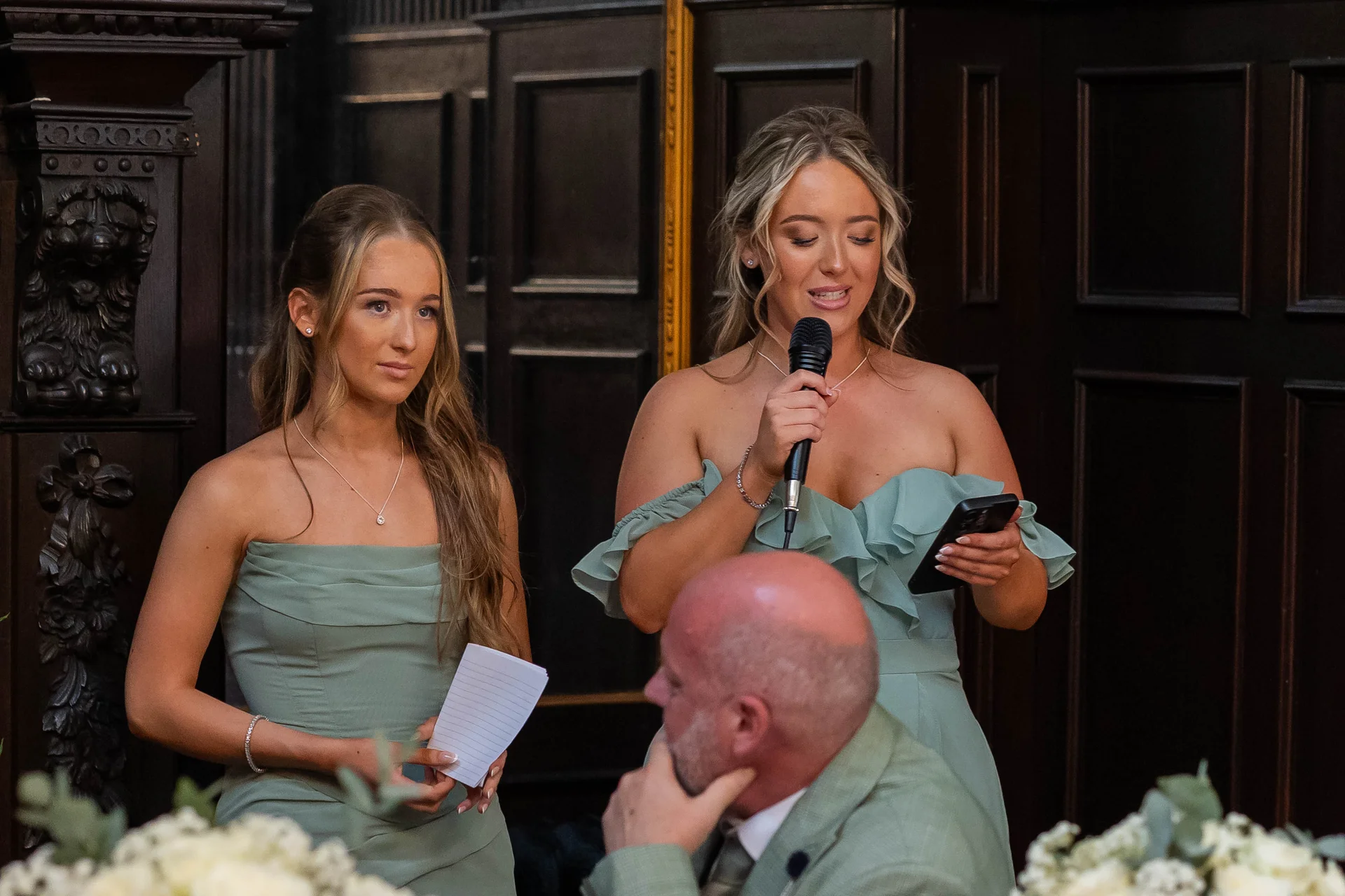 A bridesmaid delivers a heartfelt speech into a microphone while holding her phone, accompanied by another bridesmaid holding notes during the wedding reception.