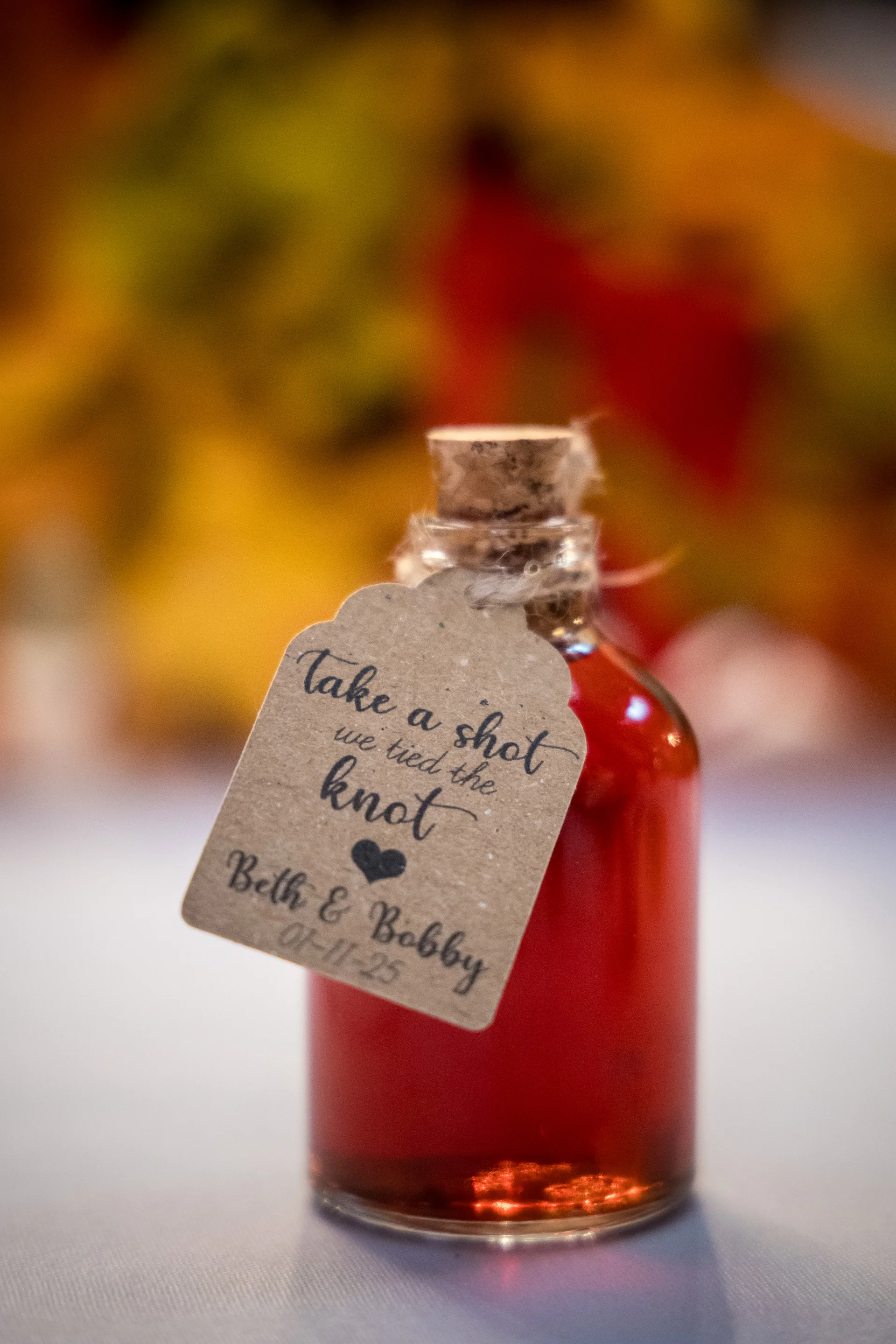 A close-up shot of a small glass wedding favour bottle filled with red liquid, featuring a rustic tag that reads take a shot we tied the knot.