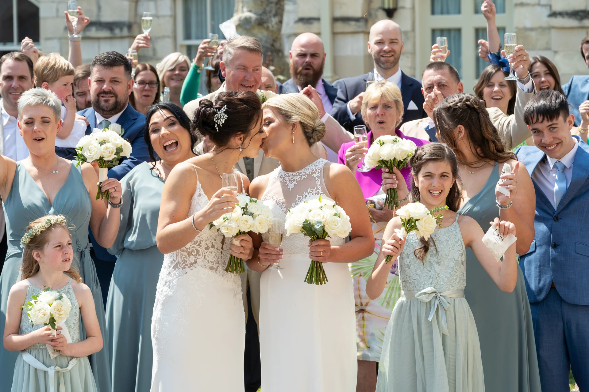 Two brides share a joyful kiss as their wedding party and guests celebrate with champagne toasts and white rose bouquets outdoors.