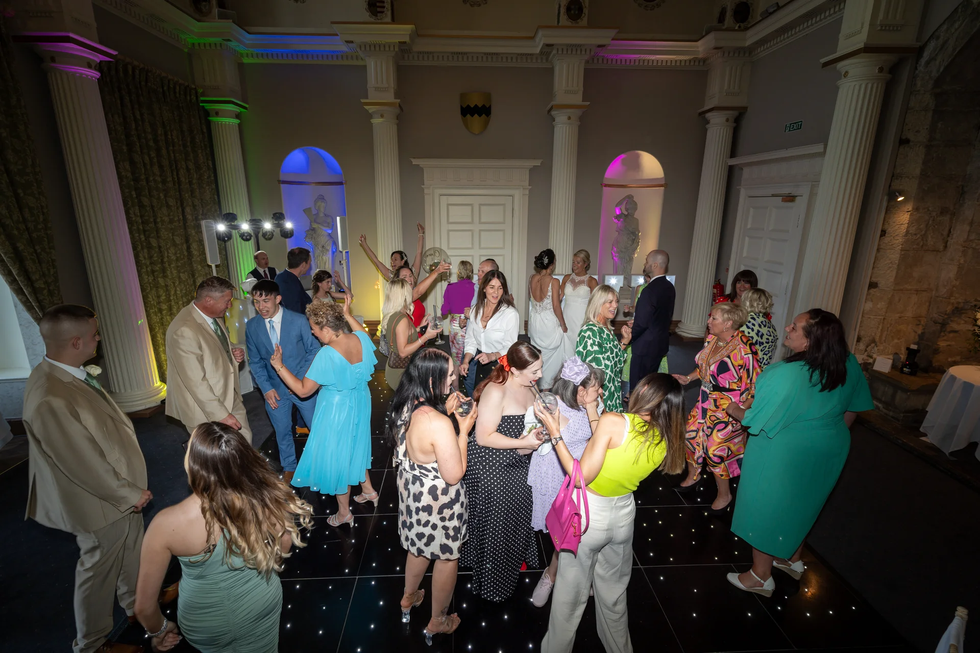 A lively evening wedding reception scene in a grand ballroom featuring guests dancing and socialising on a glowing dance floor surrounded by classical columns.