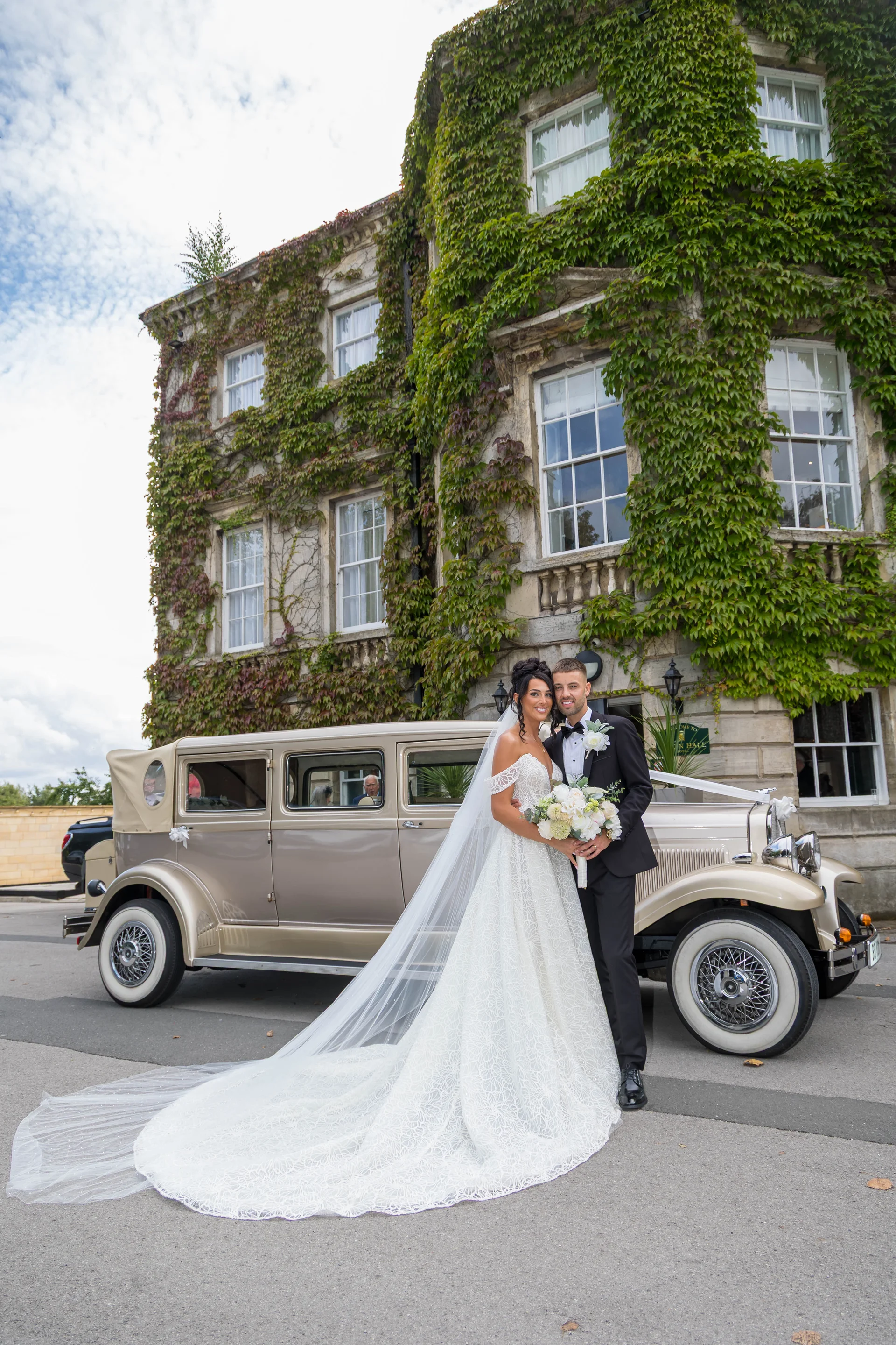 A newlywed couple poses in front of a vintage wedding car and a grand stone building covered in lush green ivy.