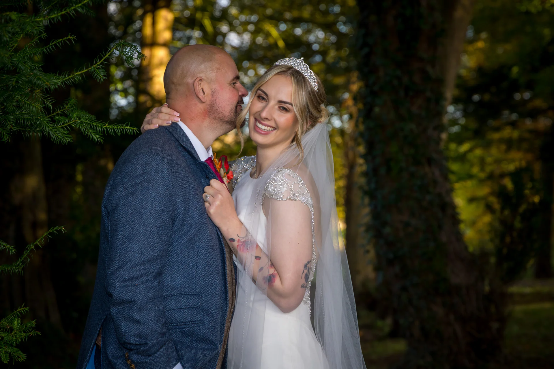 A newlywed couple embraces lovingly in a sunlit woodland setting, with the smiling bride wearing a crystal tiara and embellished gown.