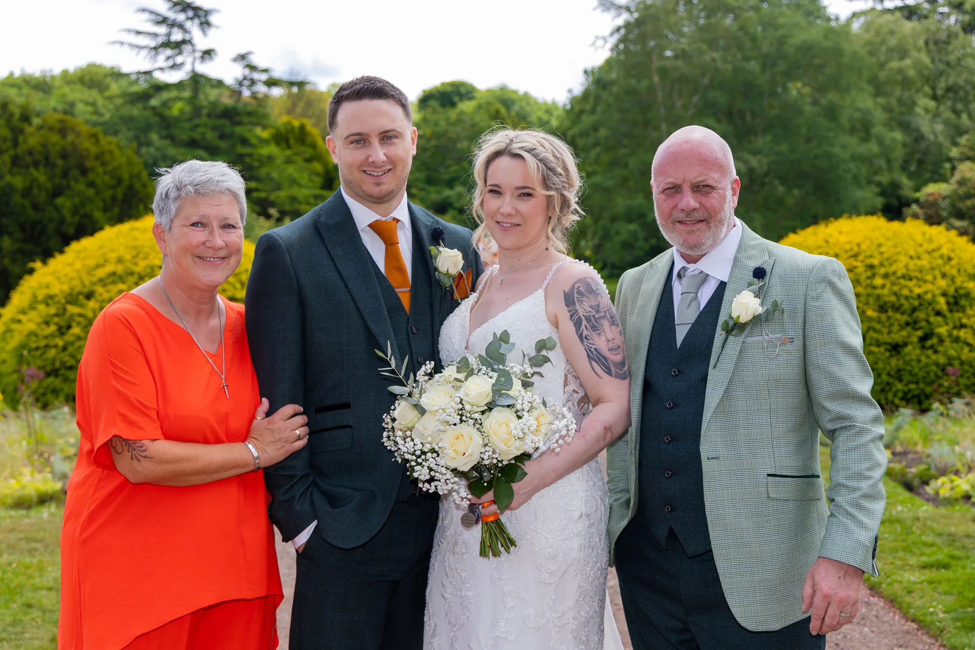 The bride and groom pose outdoors in a lush garden alongside their parents, holding a white rose bouquet.