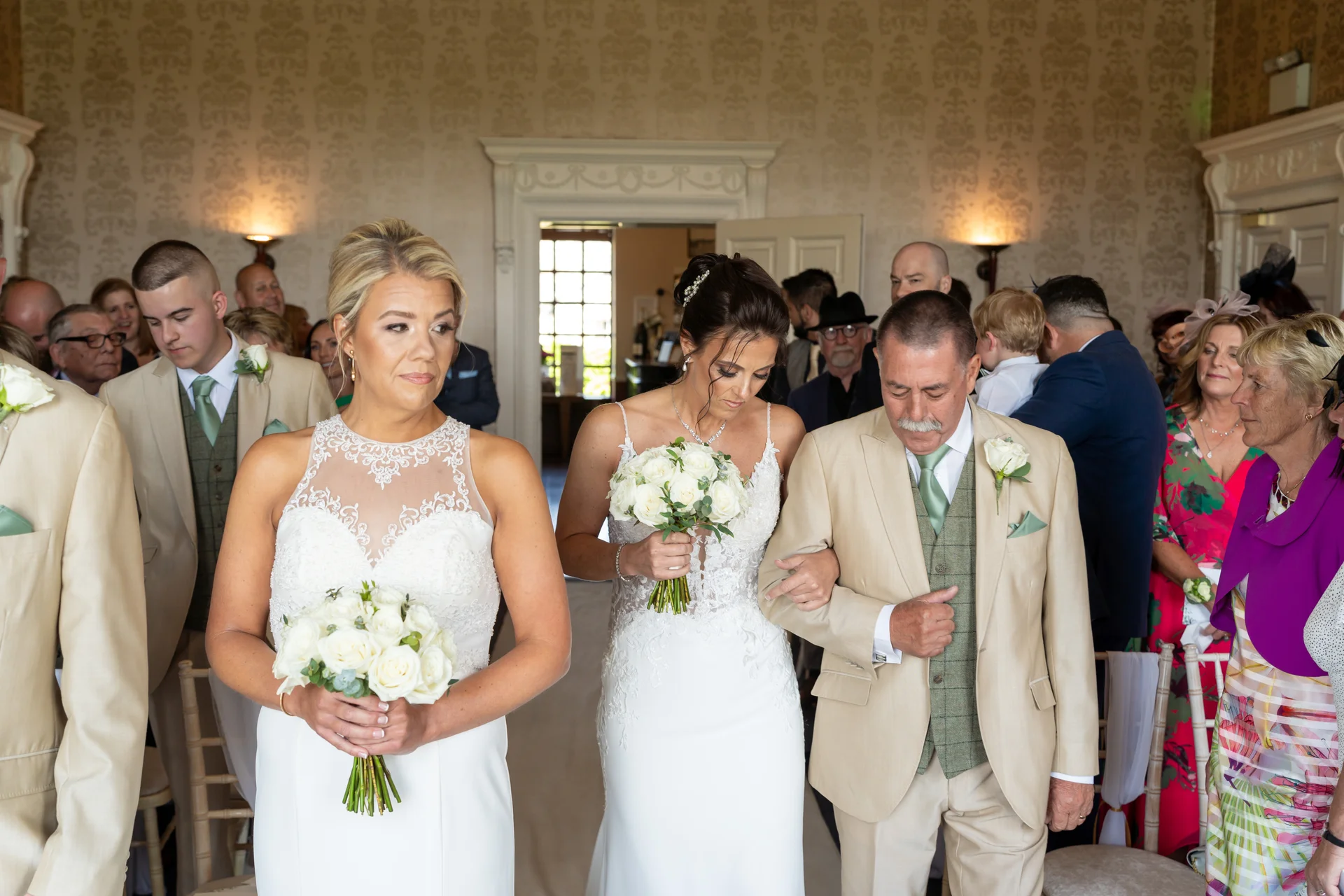 A bride walks down the aisle arm in arm with her father, preceded by a bridesmaid holding a white rose bouquet inside a grand country house venue.