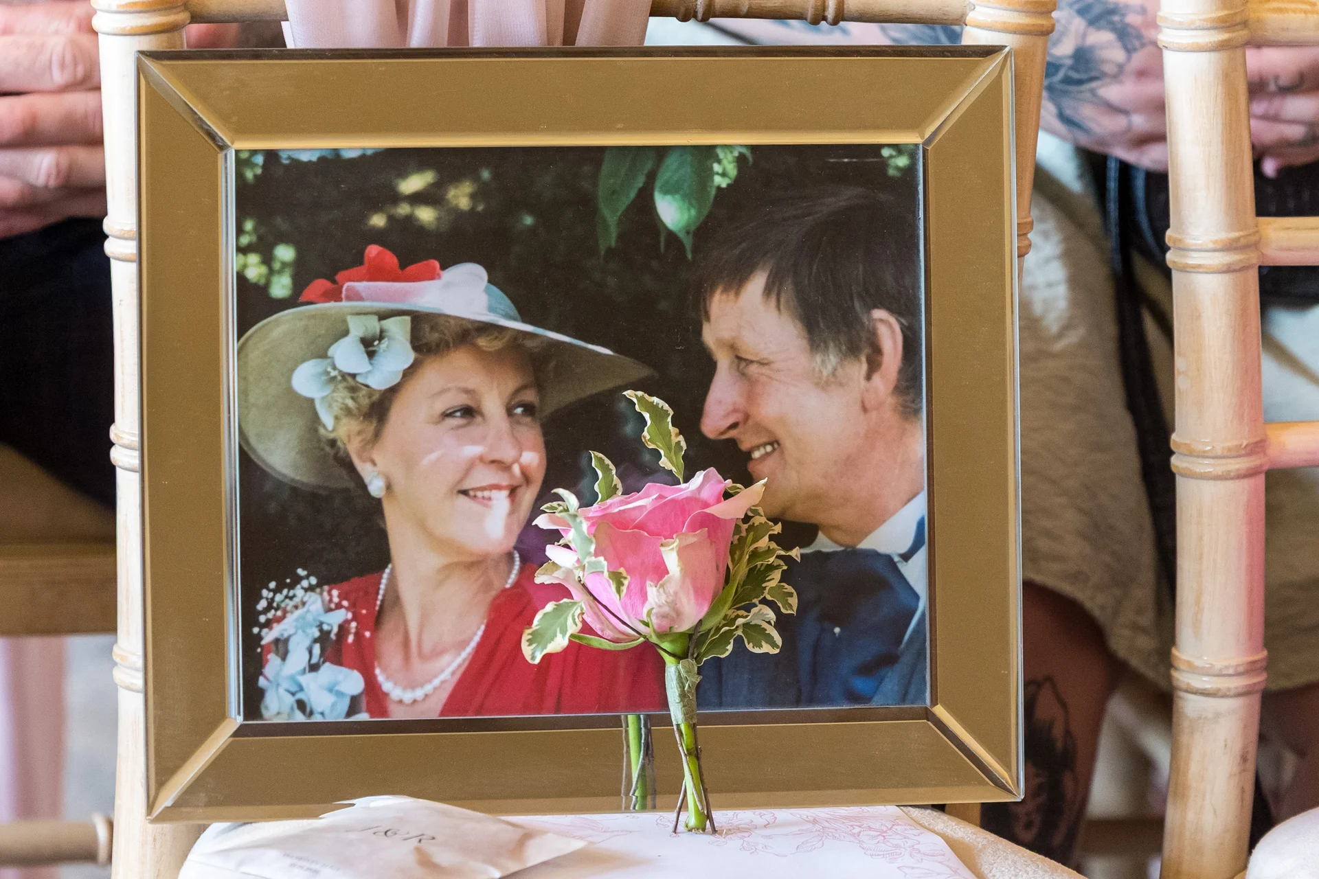 A framed vintage photograph of an older couple displayed as a memorial feature at a wedding, adorned with a single pink rose buttonhole.