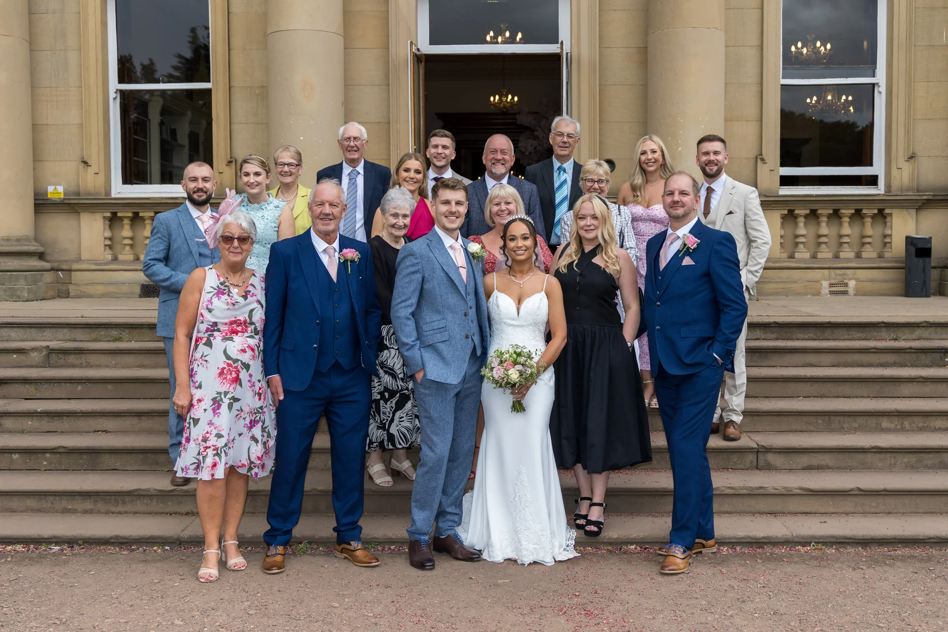 A large wedding party and family group pose together on the grand stone steps outside a stately country house venue.