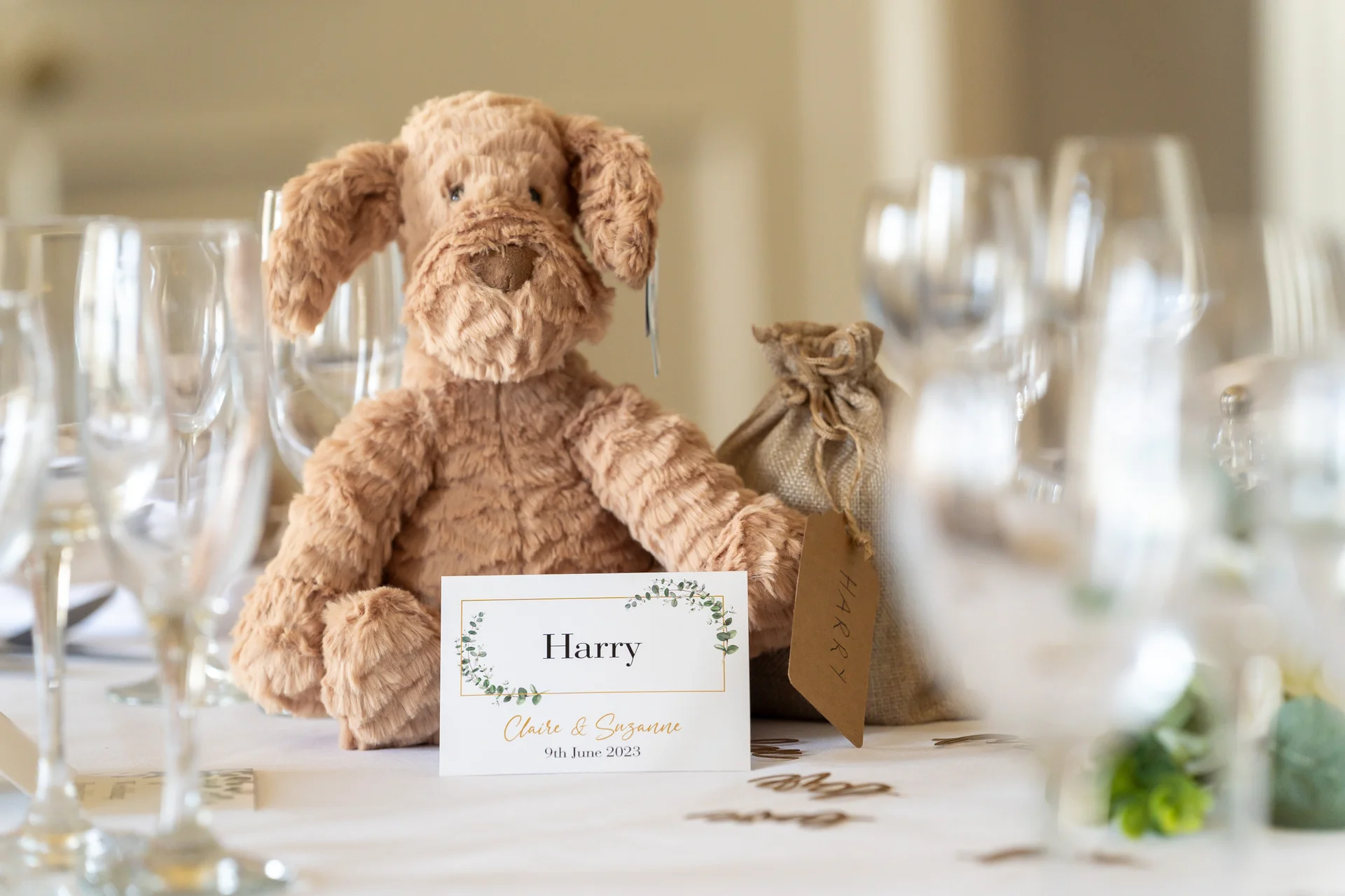 A plush stuffed dog toy sits on a wedding reception table next to a personalised place card and favour bag, surrounded by glassware.