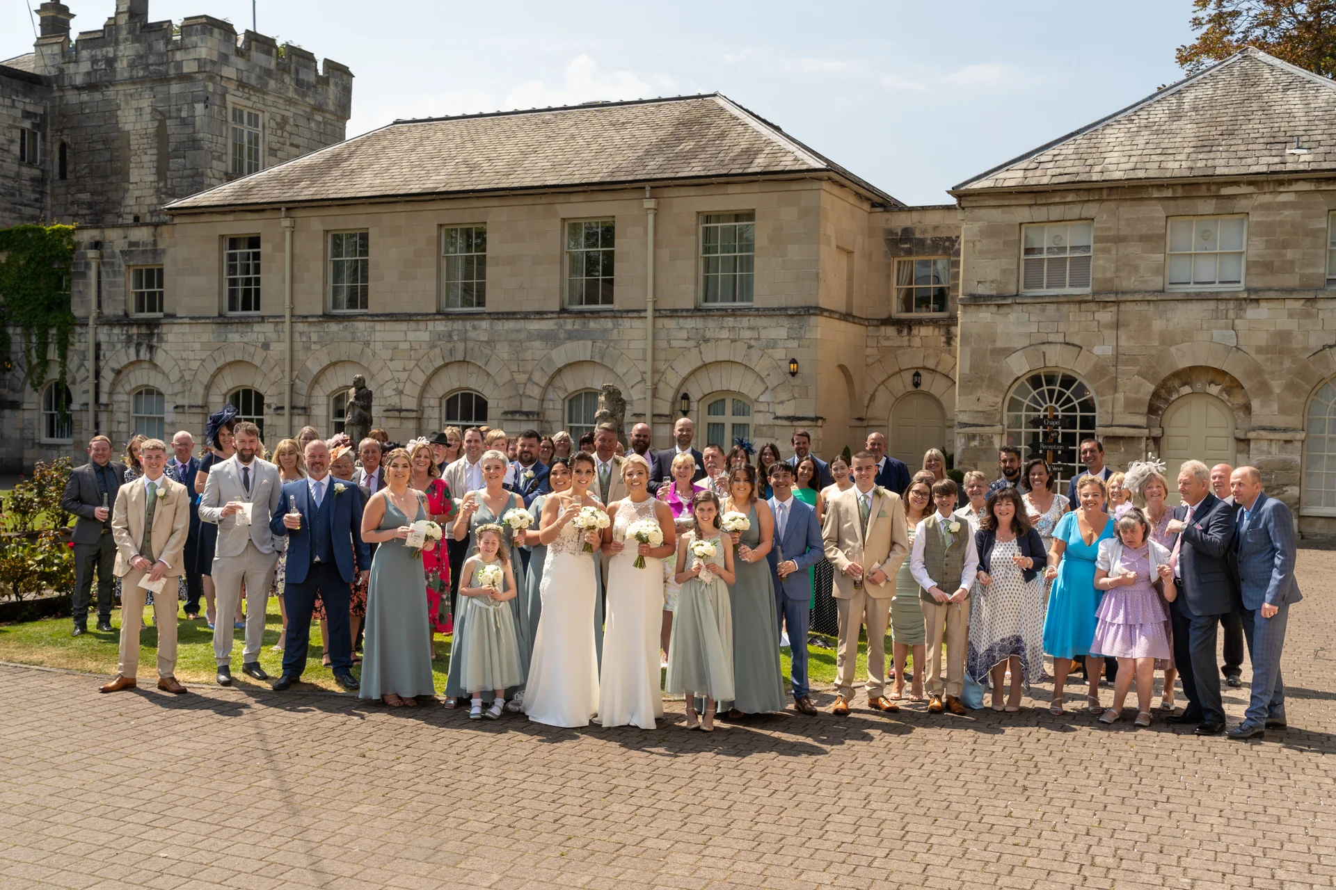 A large wedding party and guests pose for a group photo outside a historic stone castle venue on a bright sunny day.