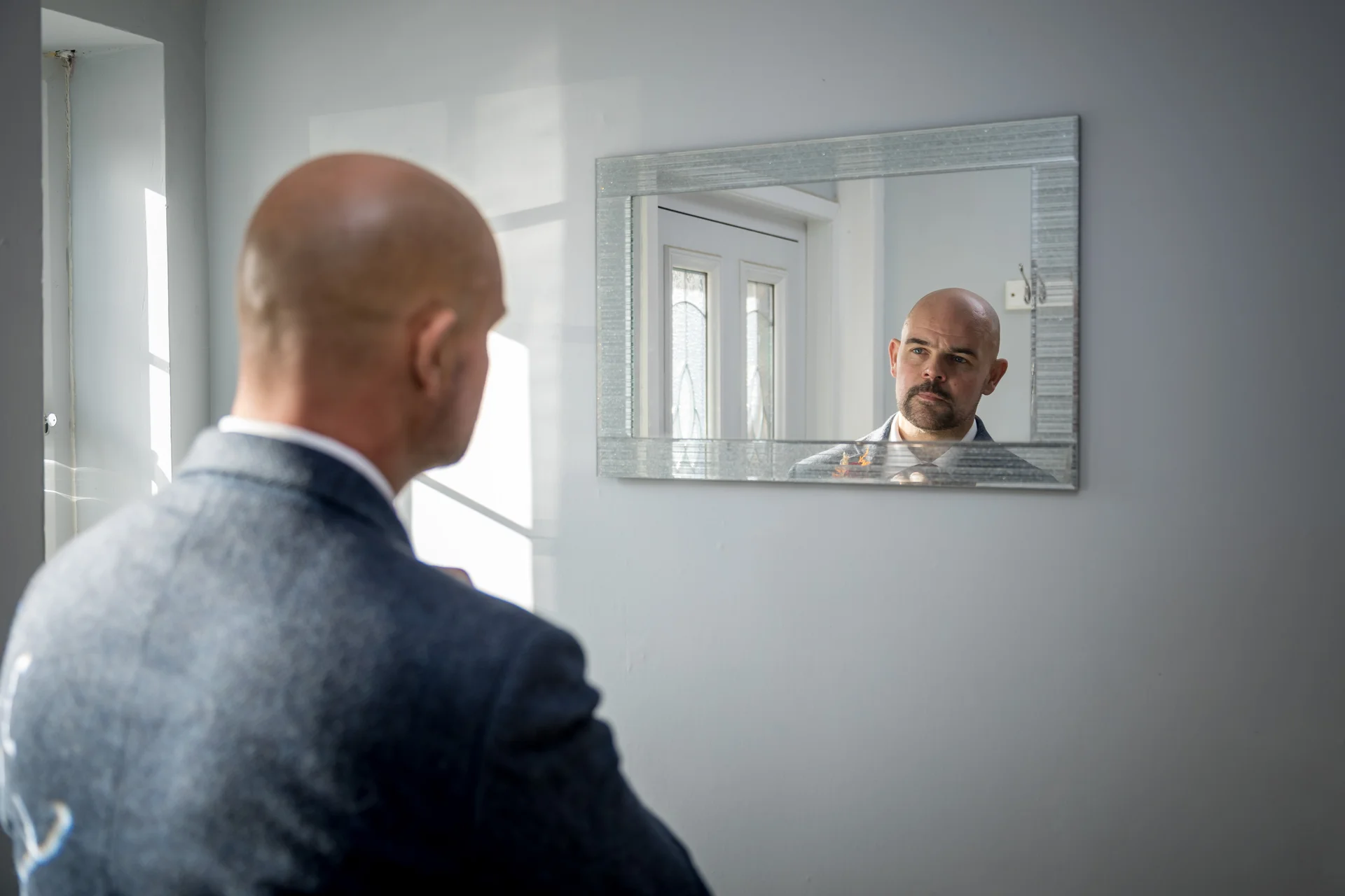 A groom stands in front of a mirror, checking his reflection and adjusting his suit during morning preparations.