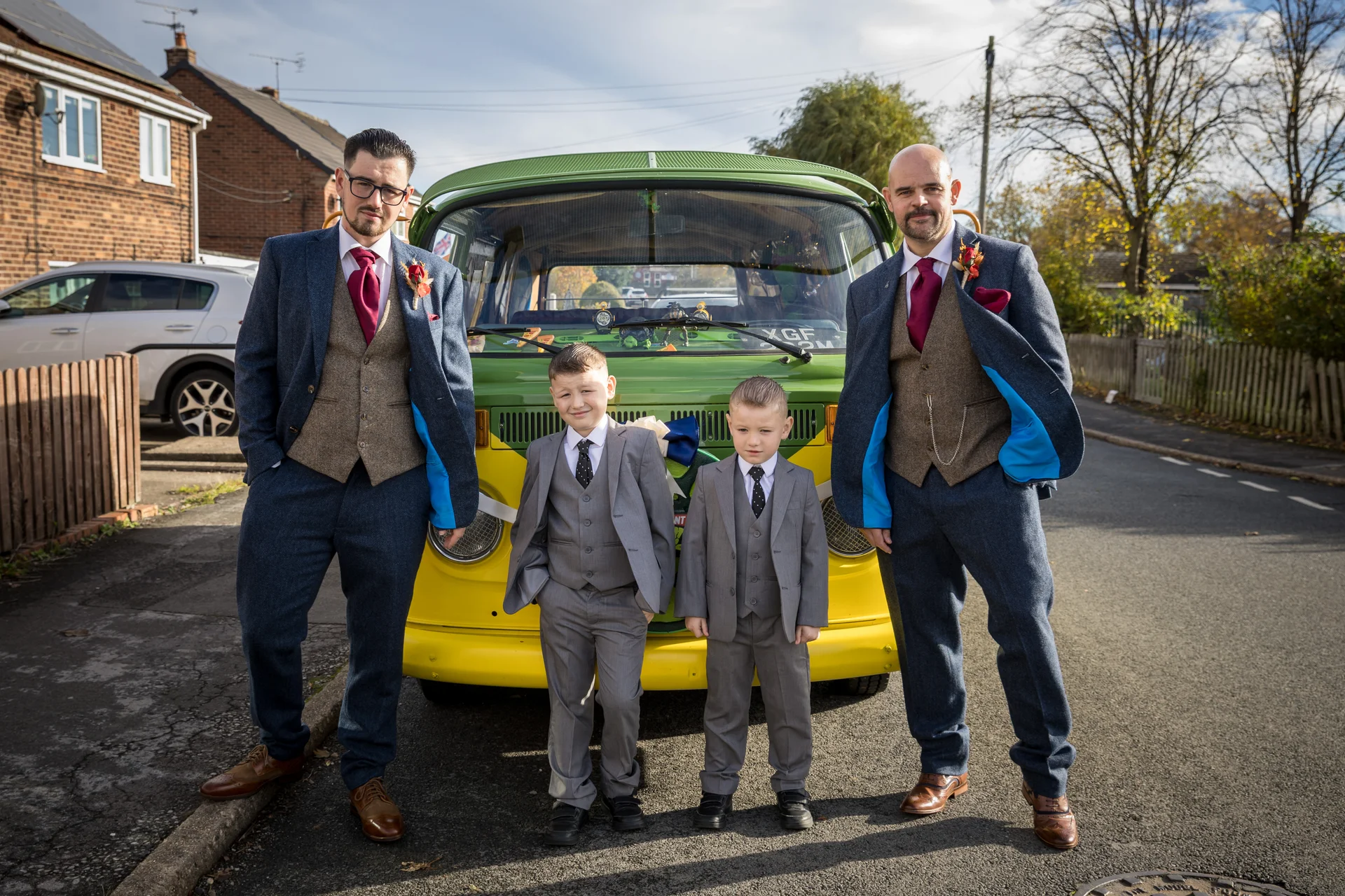 Two groomsmen in tweed suits and two page boys in grey suits pose proudly in front of a vintage green and yellow campervan on a sunny street.