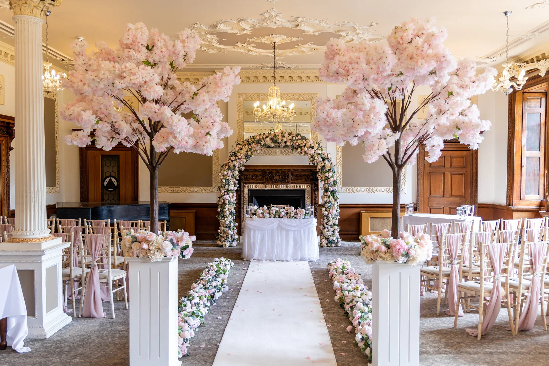 A grand ballroom styled for a wedding ceremony features an aisle lined with pink blossom trees, floral runners, and chairs draped with pink sashes leading to a floral arch.