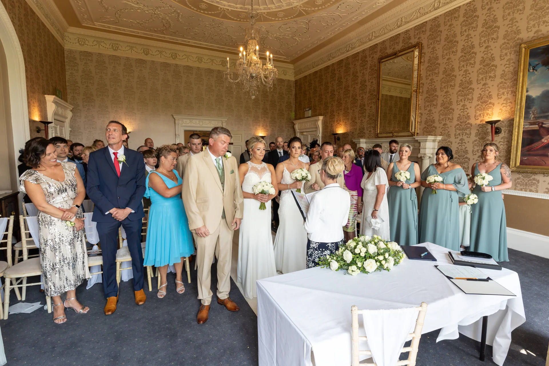 A civil marriage ceremony takes place in an elegant, panelled country house room, featuring two brides standing side by side before the celebrant.