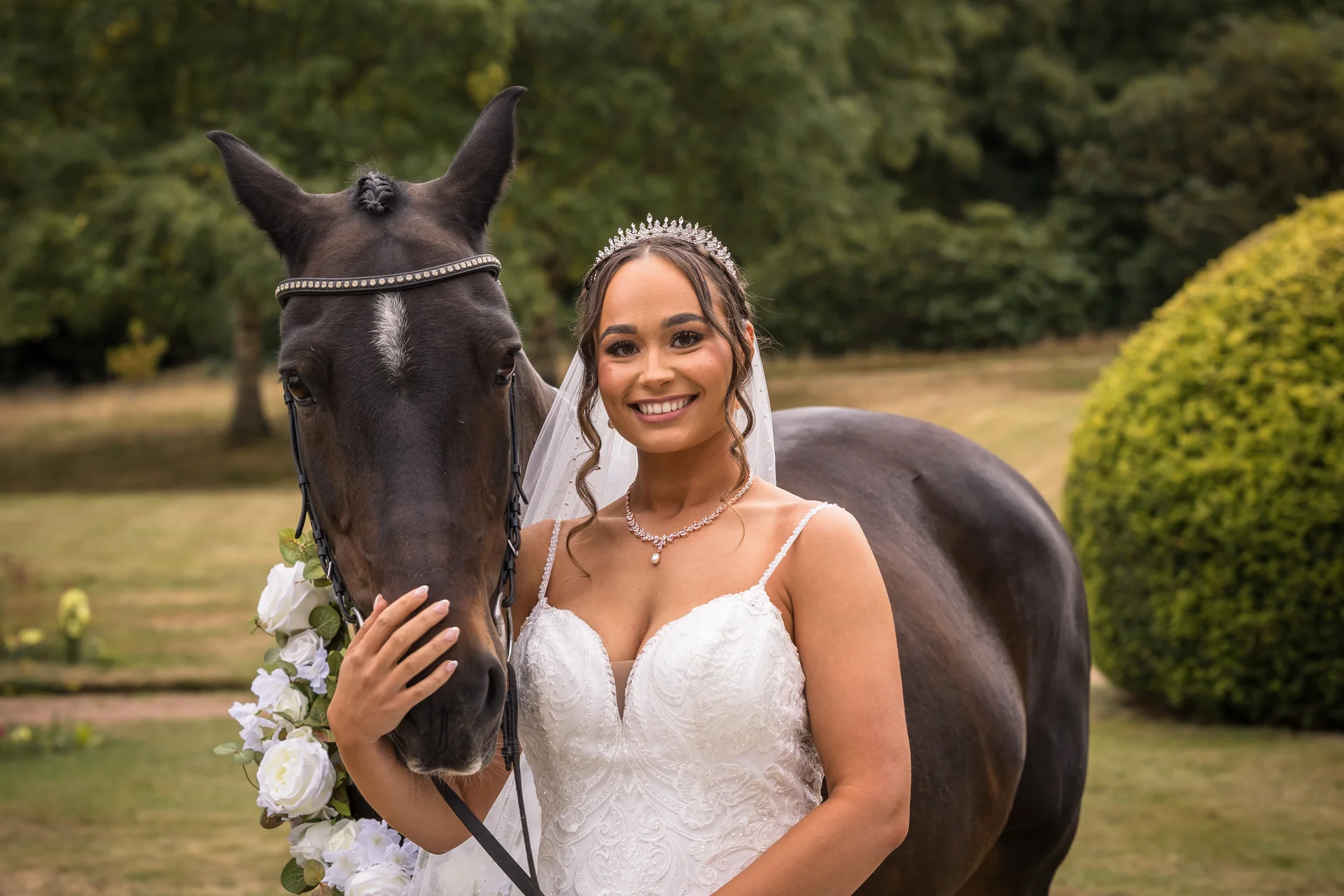 A smiling bride wearing a crystal tiara and lace gown poses outdoors next to a dark horse adorned with white floral garlands.