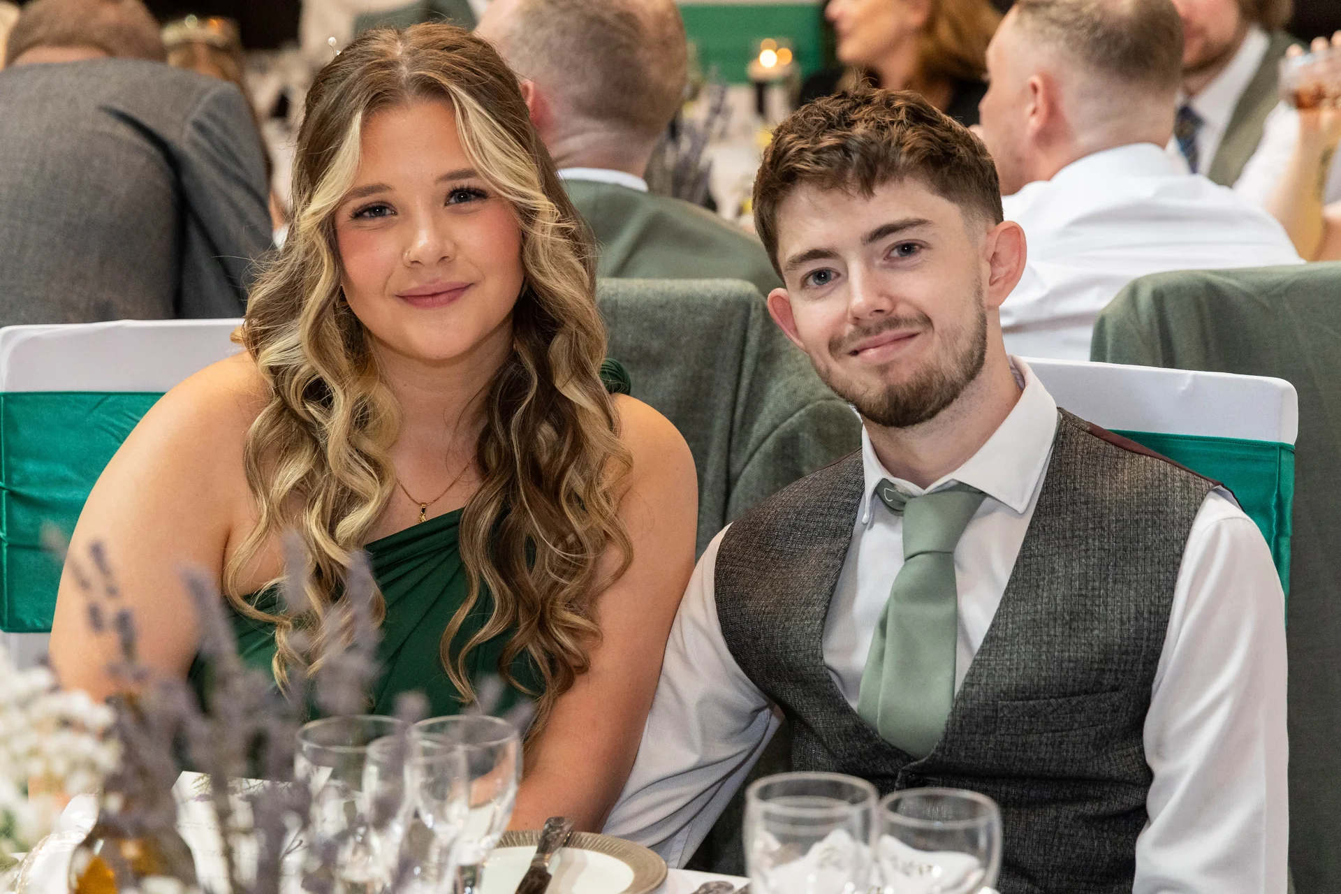 A guest and her partner pose together smiling at a wedding reception table adorned with glassware and lavender decor.