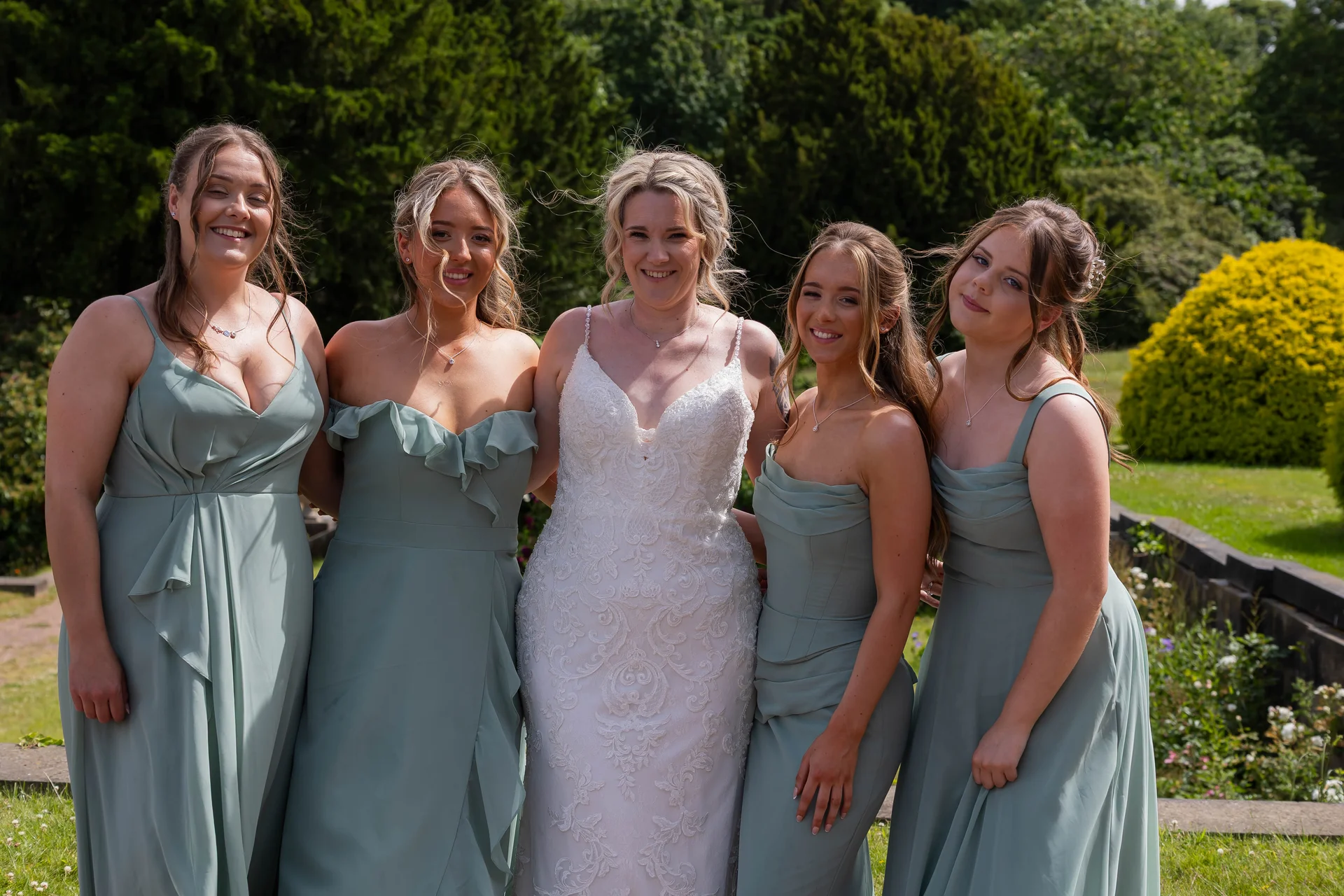 The radiant bride in a lace gown stands outdoors in a sunny garden flanked by four smiling bridesmaids wearing matching sage green dresses.