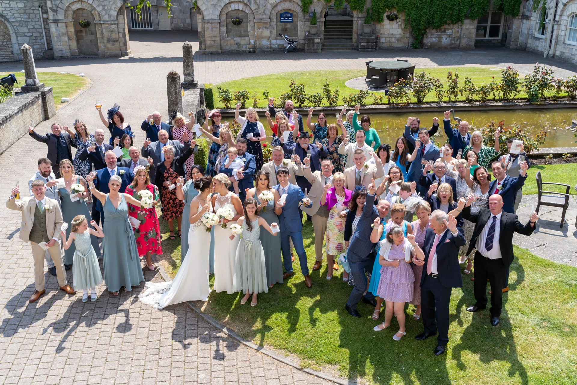 A large group of wedding guests cheer and raise glasses during a celebration in a sunny stone castle courtyard featuring a manicured lawn and ornamental pond.