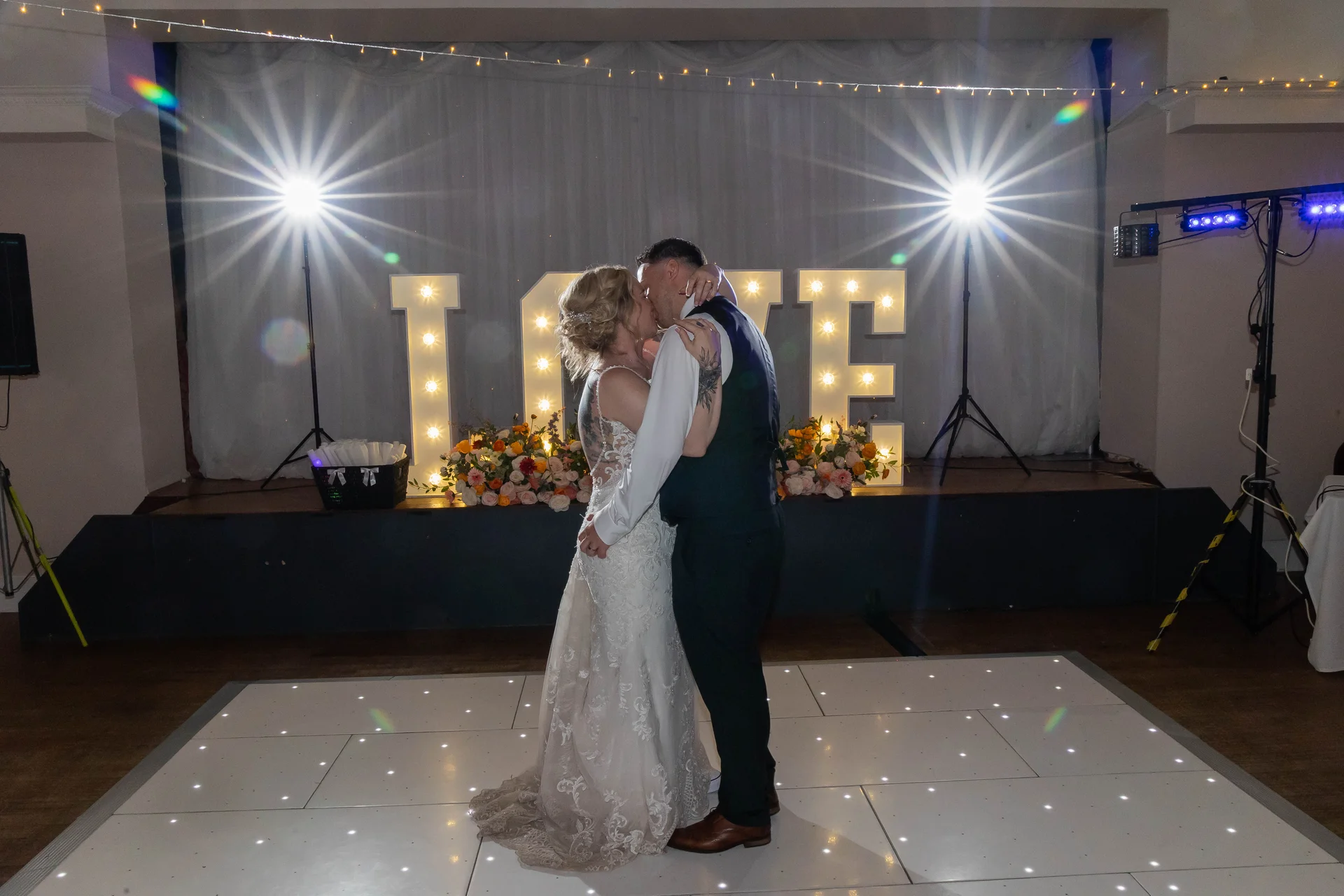 Newlyweds share a tender kiss during their first dance on an illuminated starlight dance floor in front of large glowing love letters.