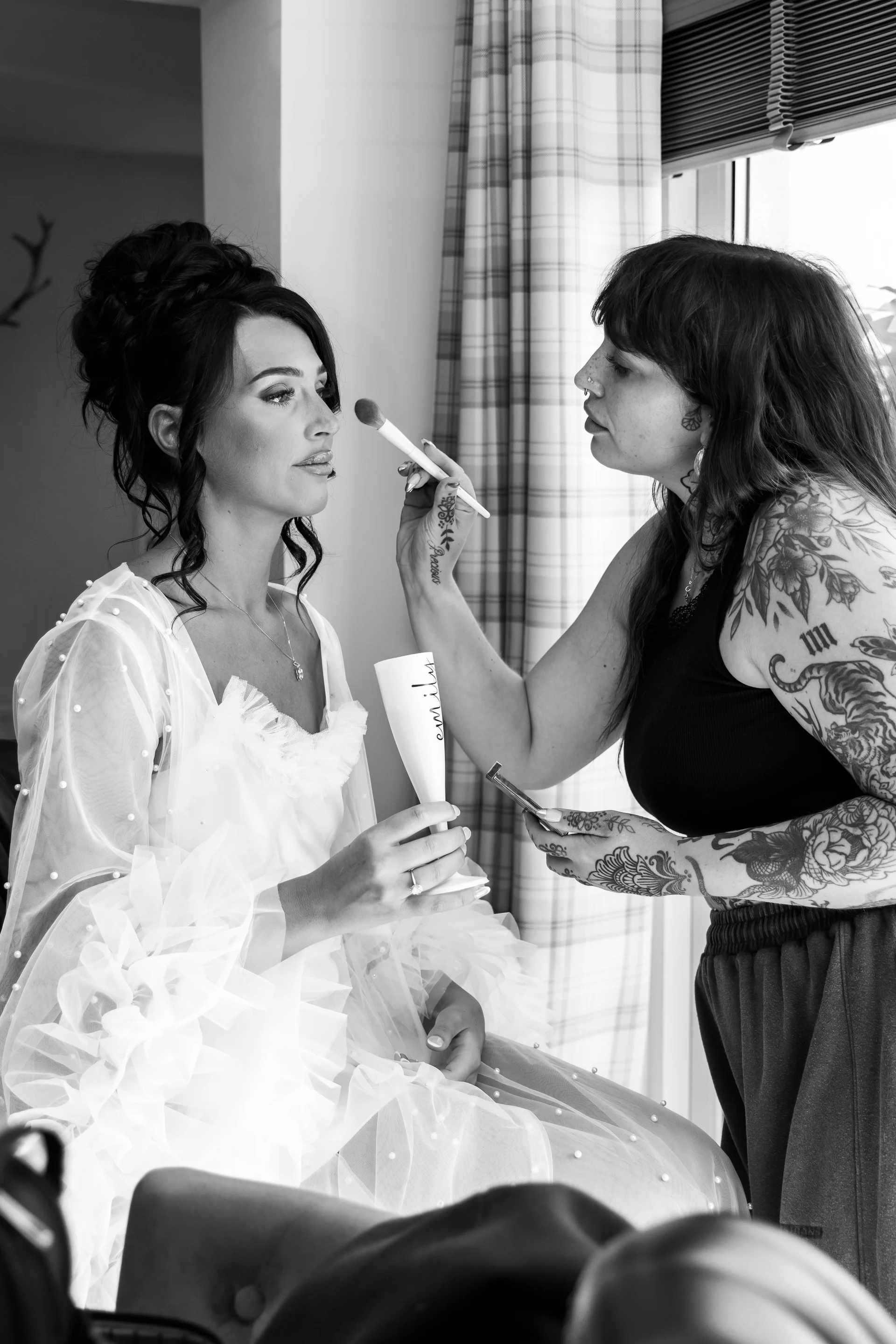 A black and white photograph captures a bride having her makeup applied by a professional artist near a bright window while holding a personalised champagne glass during morning preparations.
