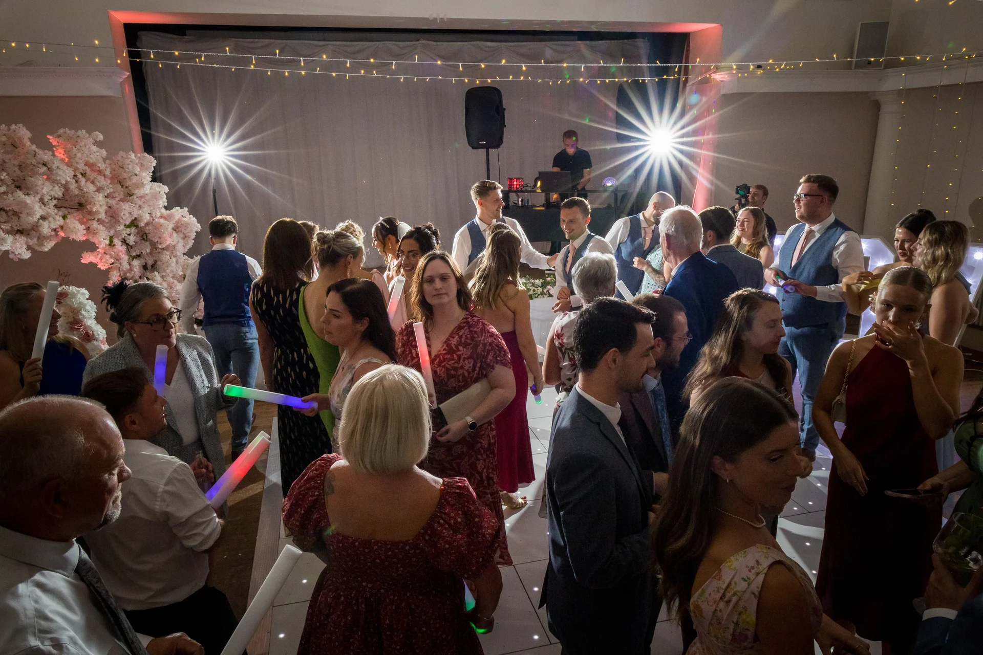 A lively wedding evening reception featuring guests mingling and holding glowing foam sticks on a illuminated dance floor under festive string lights.