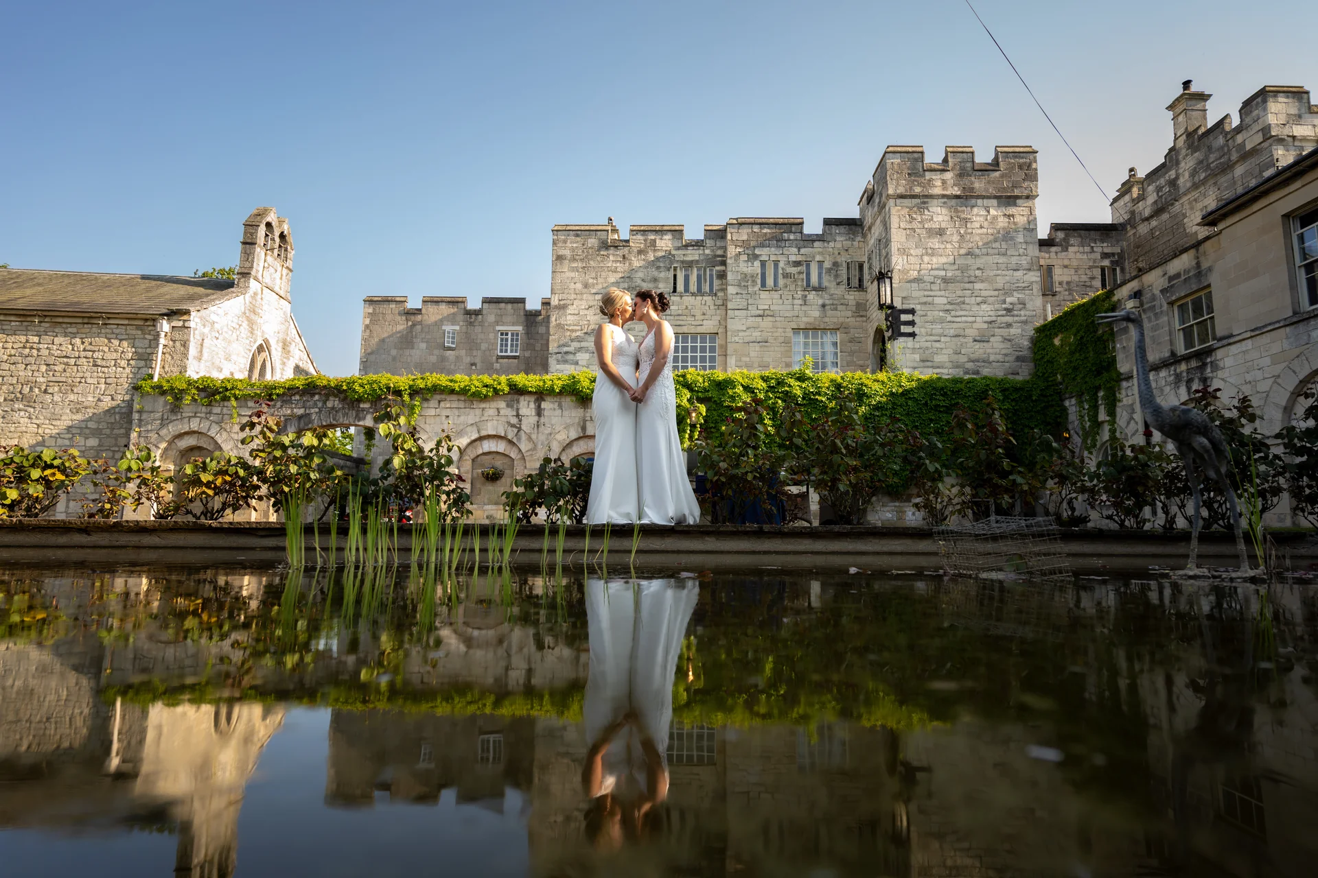Two brides stand hand in hand sharing a romantic moment outside a historic stone castle, perfectly mirrored in the tranquil pond in the foreground.