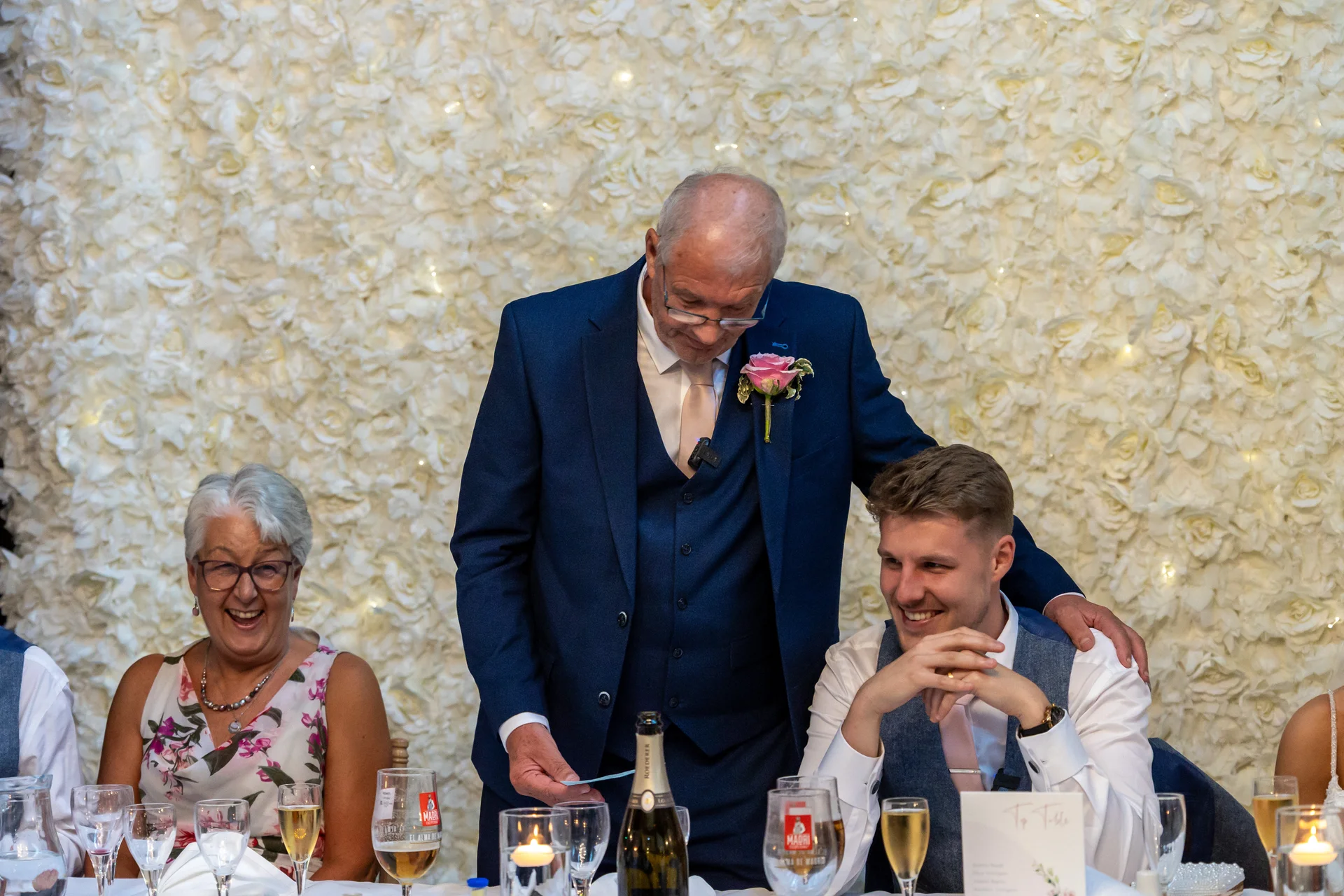 An older man in a navy suit gives a speech at the top table in front of a large white rose flower wall while family members laugh and smile.