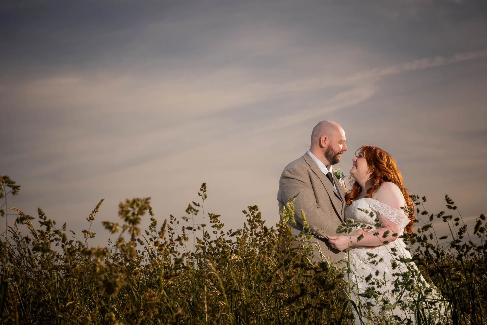 A newlywed couple embrace lovingly amongst tall meadow grass beneath a warm sunset sky during their golden hour portrait session.