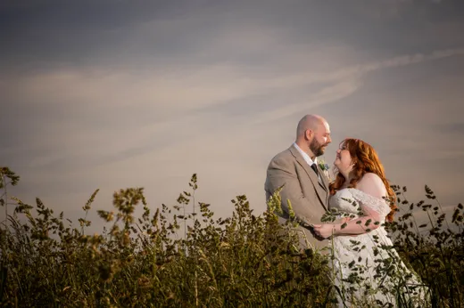 A newlywed couple embrace lovingly amongst tall meadow grass beneath a warm sunset sky during their golden hour portrait session.