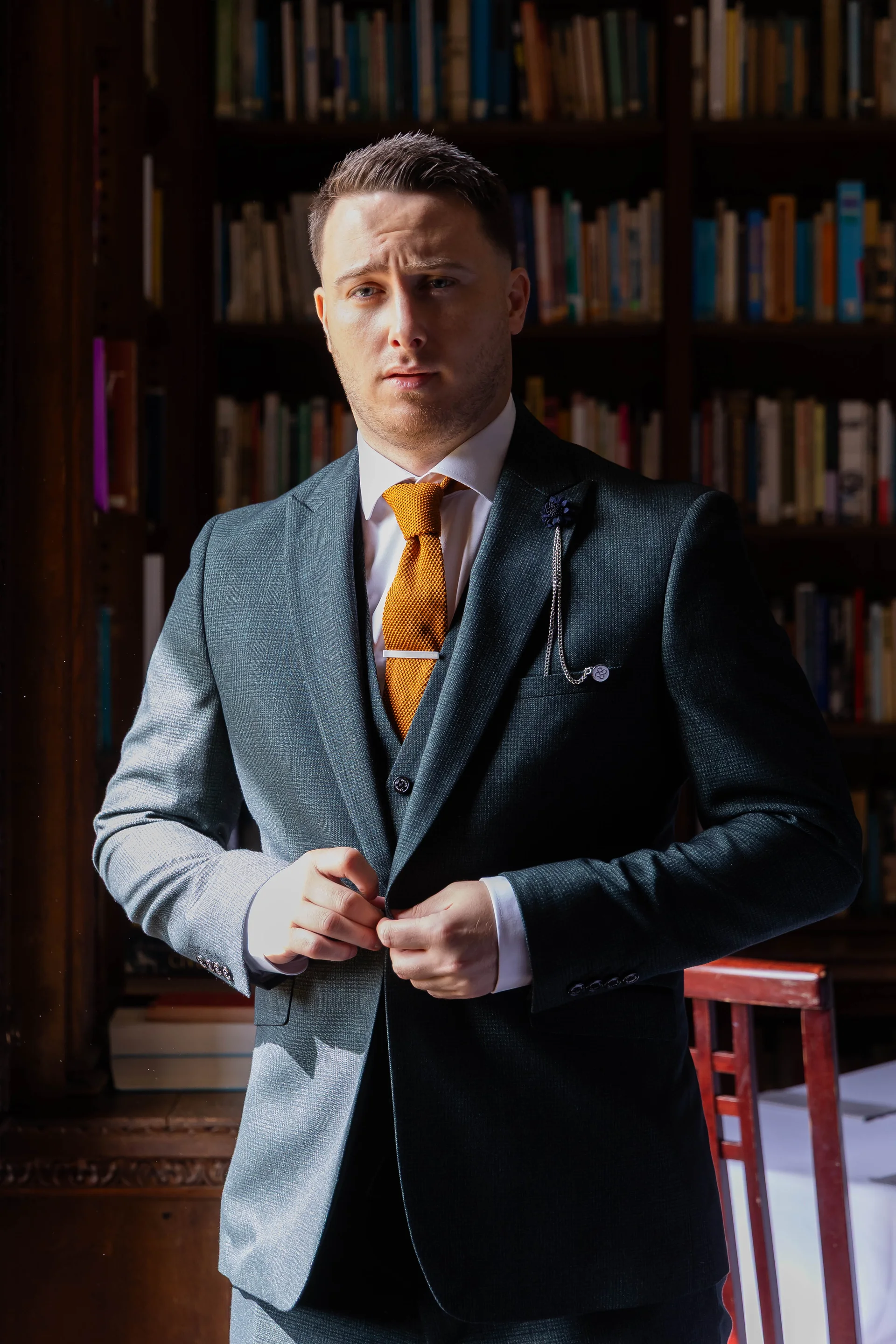 A handsome groom in a dark grey three-piece suit adjusts his jacket inside a classic library with floor-to-ceiling bookshelves.