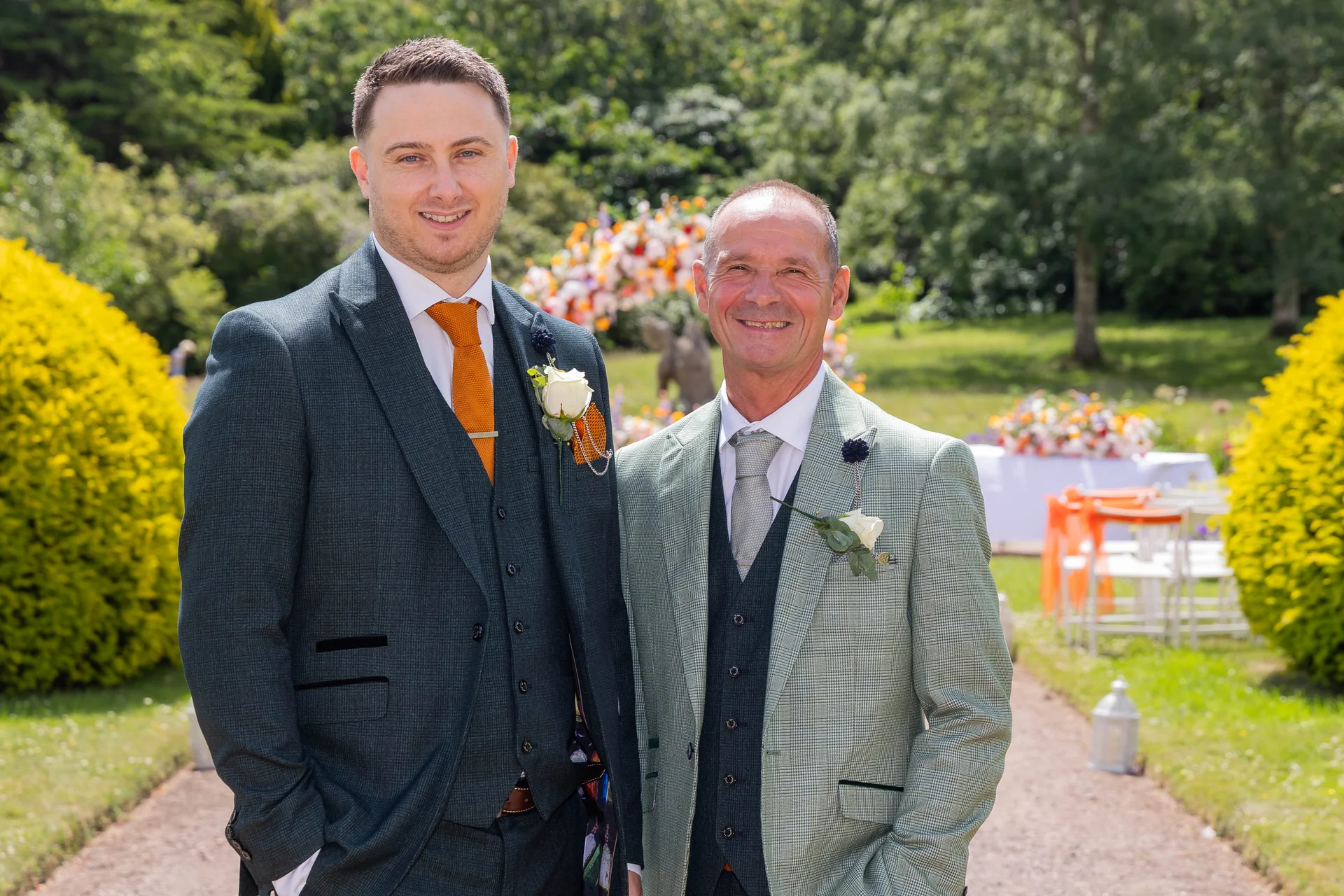 The groom and an older family member stand smiling together outdoors in a sunlit garden wearing tailored suits with boutonniere floral lapel pins.