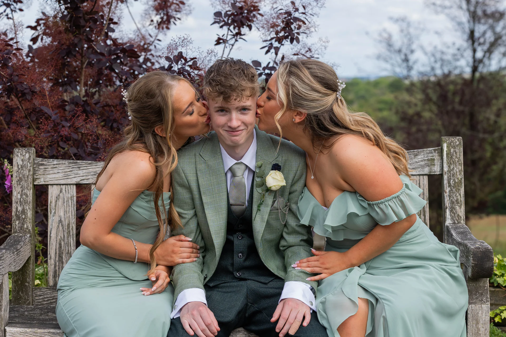 Two bridesmaids in sage green dresses playfully kiss a young man in a tweed suit seated on a wooden garden bench.