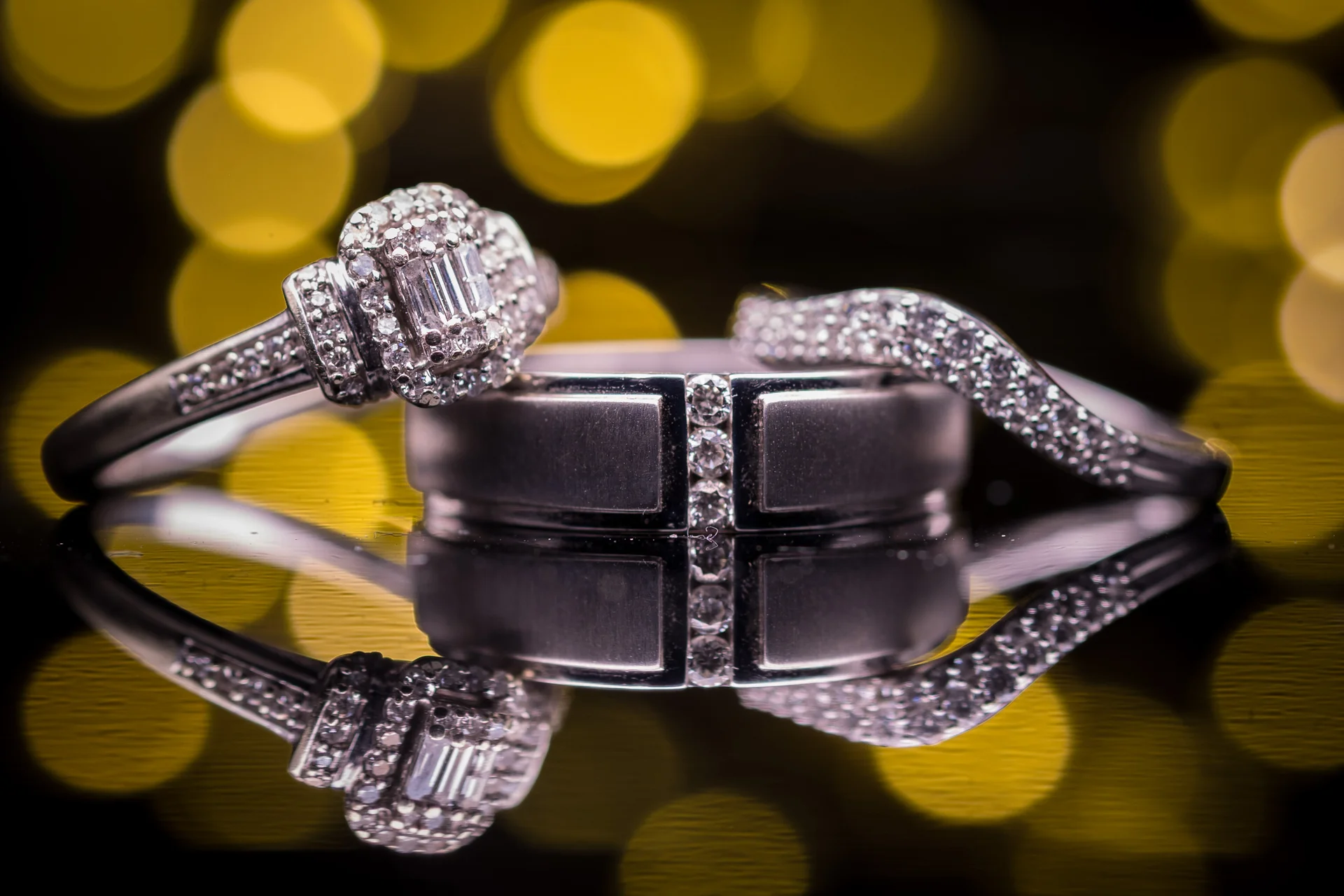 A macro detail shot showcases three diamond-embellished wedding rings arranged on a reflective surface against a warm, glowing bokeh background.
