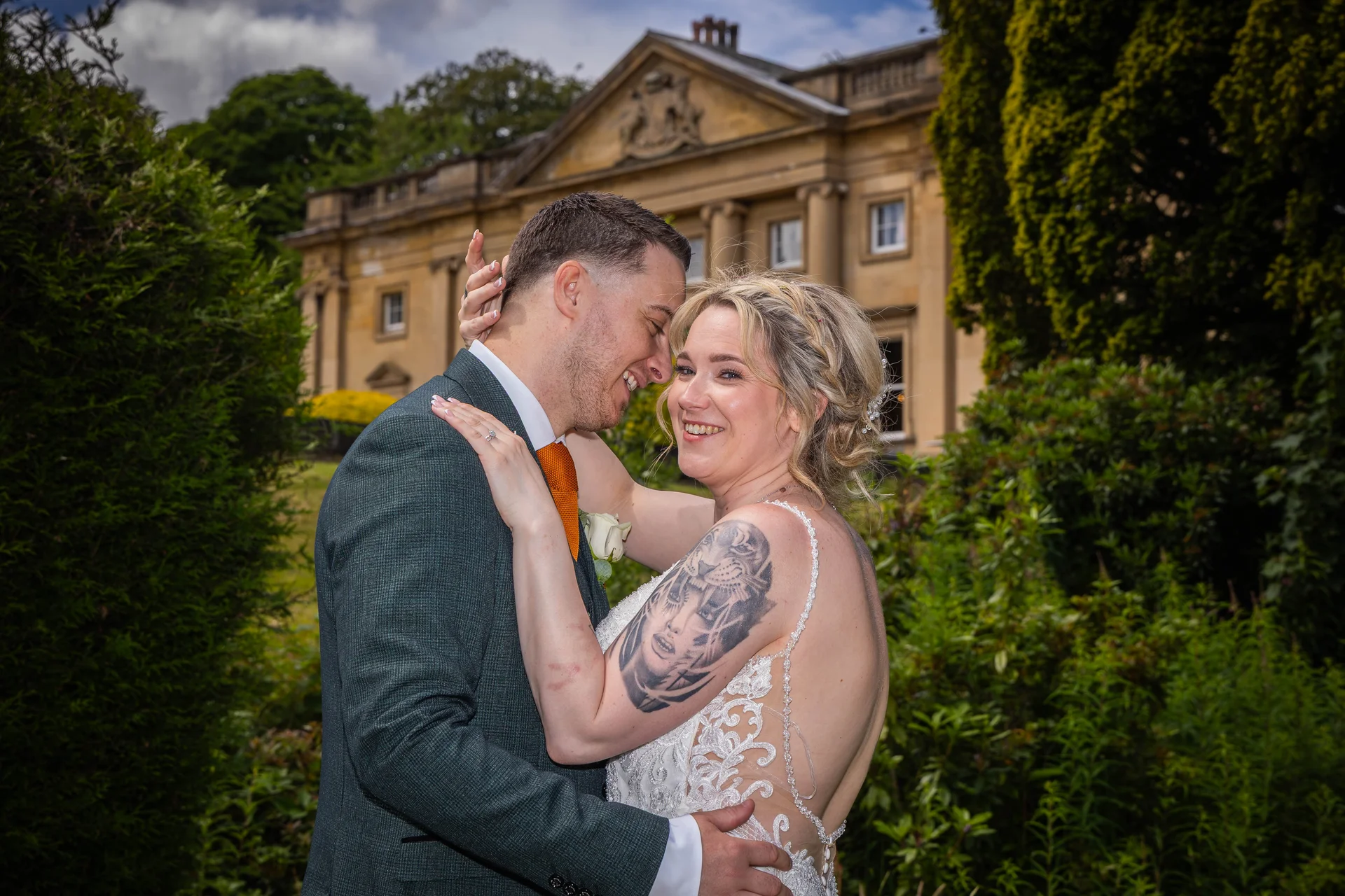 A newlywed couple embraces and laughs together in the manicured gardens of a stately country house venue.