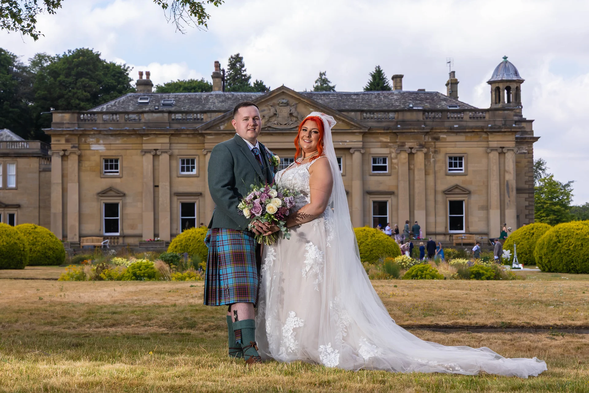 A newlywed couple poses on the grounds of a grand country house, with the groom wearing a traditional tartan kilt and the bride in a lace gown with a long veil.
