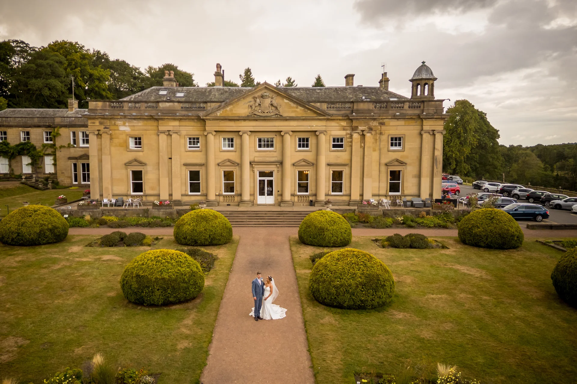 A newlywed couple stands on a pathway in front of a grand manor house, viewed from above with manicured gardens and topiary bushes.