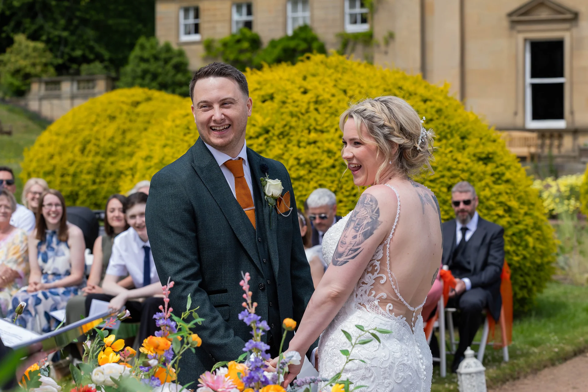 A joyful bride and groom laugh together during an outdoor ceremony at a grand country house, surrounded by seated guests and vibrant floral arrangements.