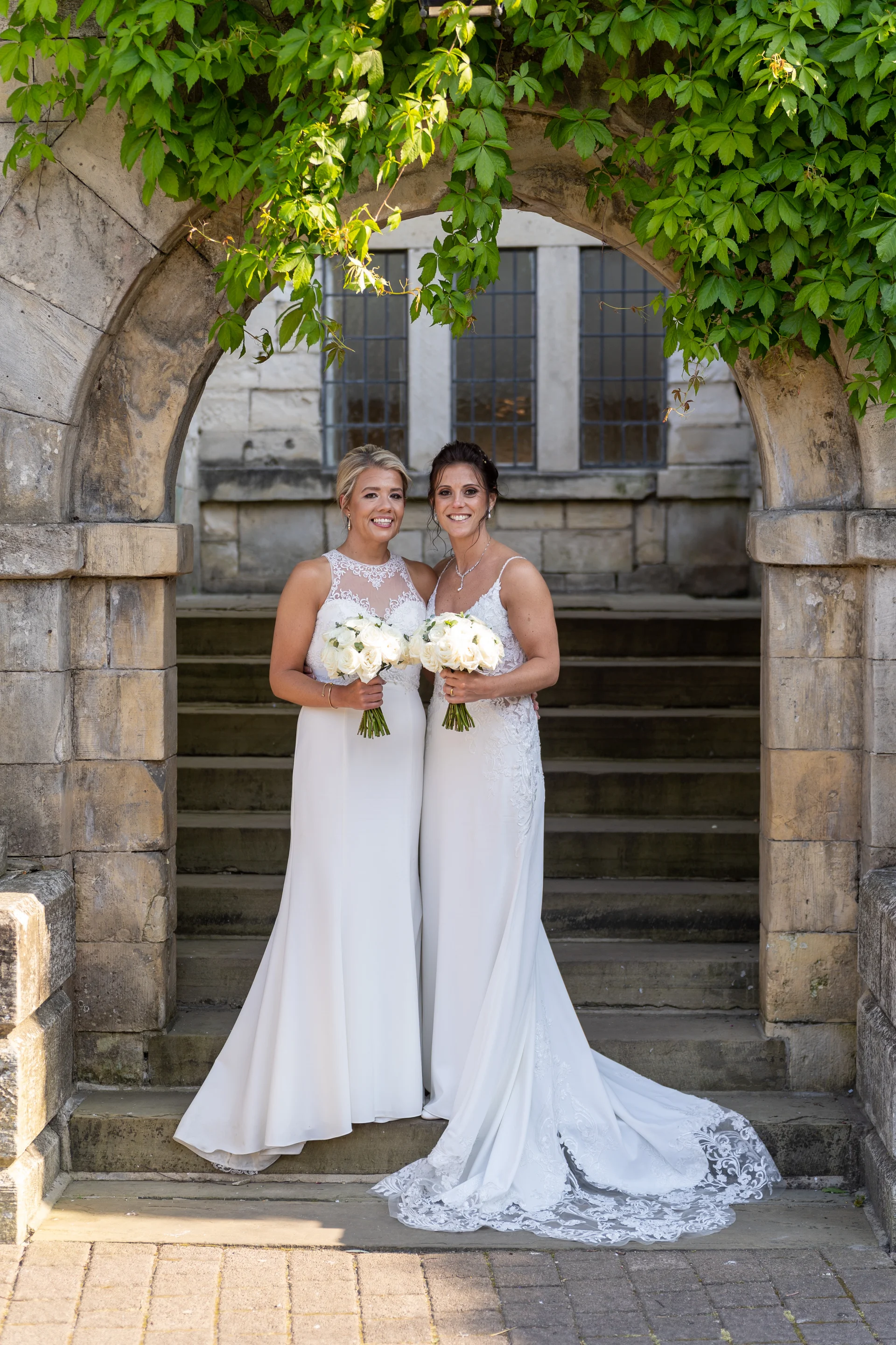 Two smiling brides stand side by side holding bouquets of white roses, framed within an ivy-covered stone archway.