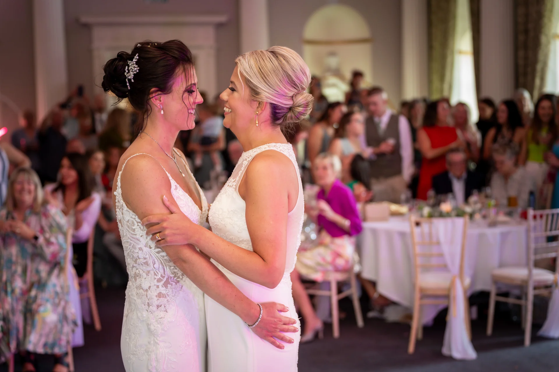 Two brides share a tender moment dancing together during their wedding reception while guests watch and mingle in the background.